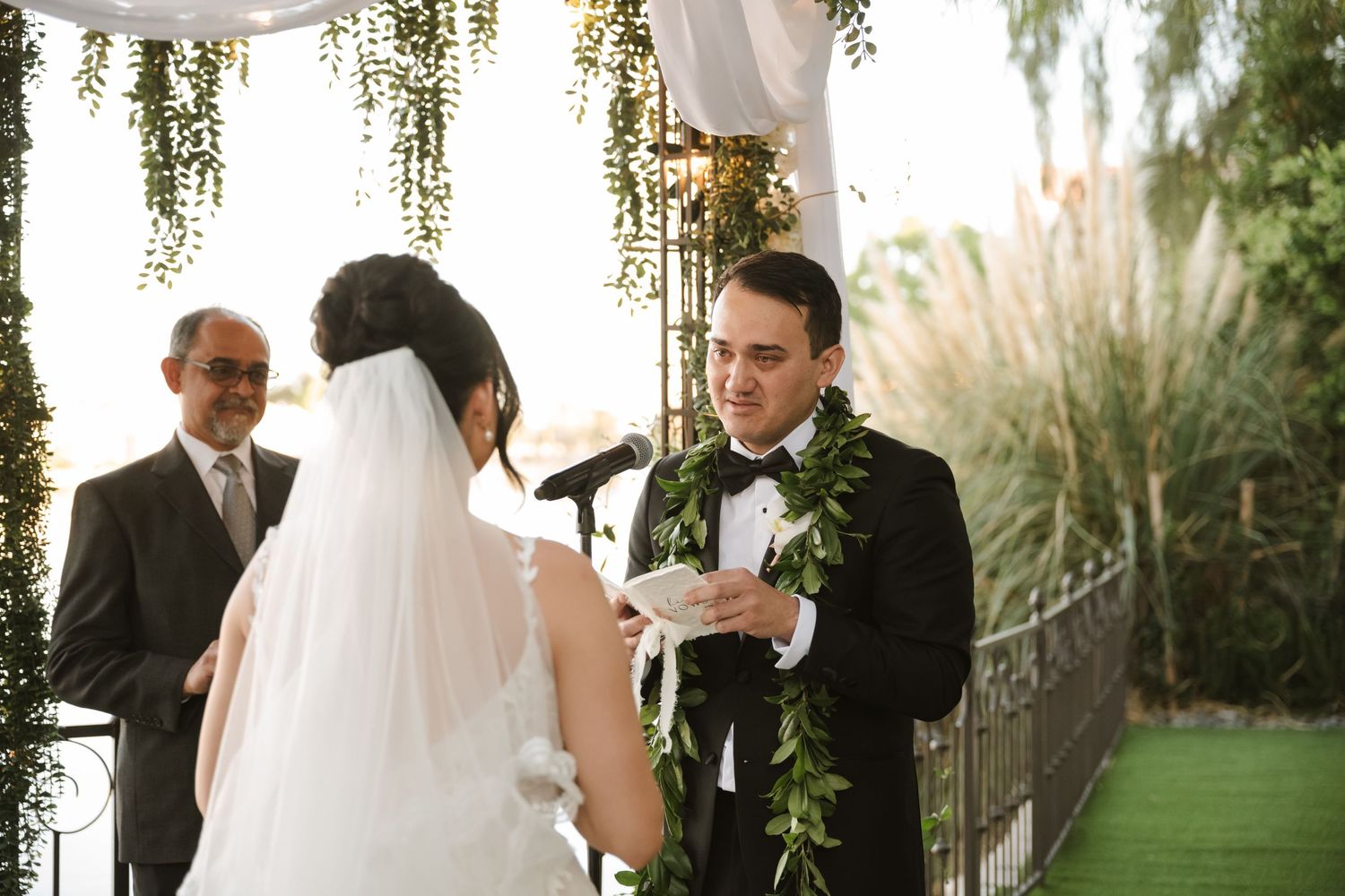 Groom in black tuxedo with floral lei reads wedding vows to bride in white dress at outdoor ceremony.