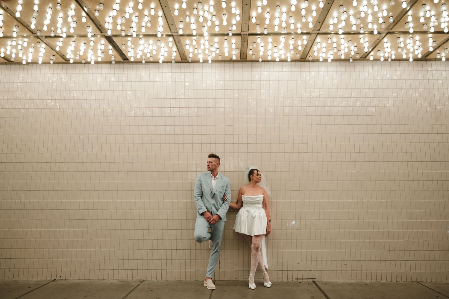 Stylish couple posing against a tiled wall with string lights, bride in short white dress and groom in light blue suit.