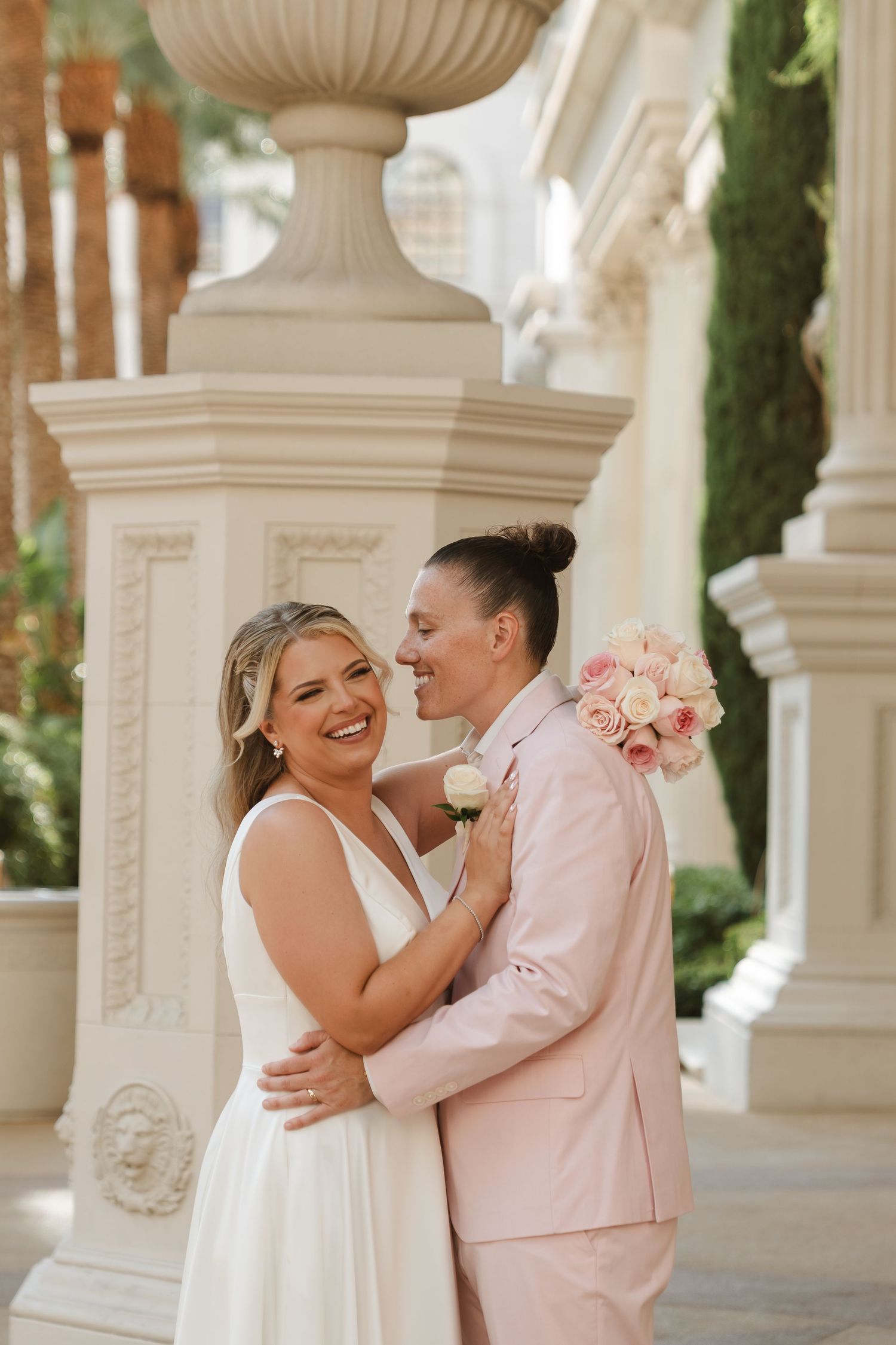 Couple in white and pink attire laughing together under ornate white columns.