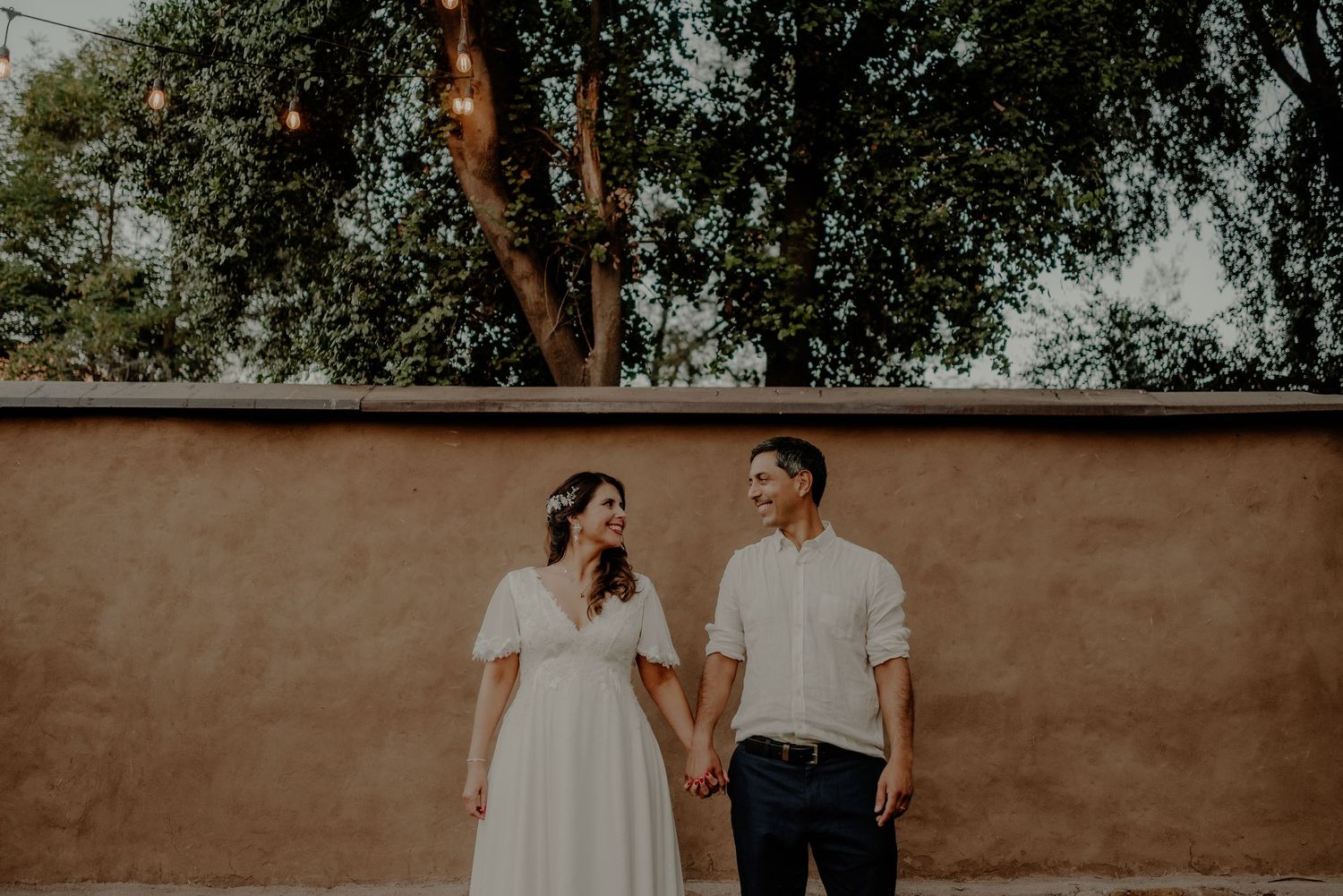 A casual engagement photo of a couple against a rustic wall in natural lighting.
