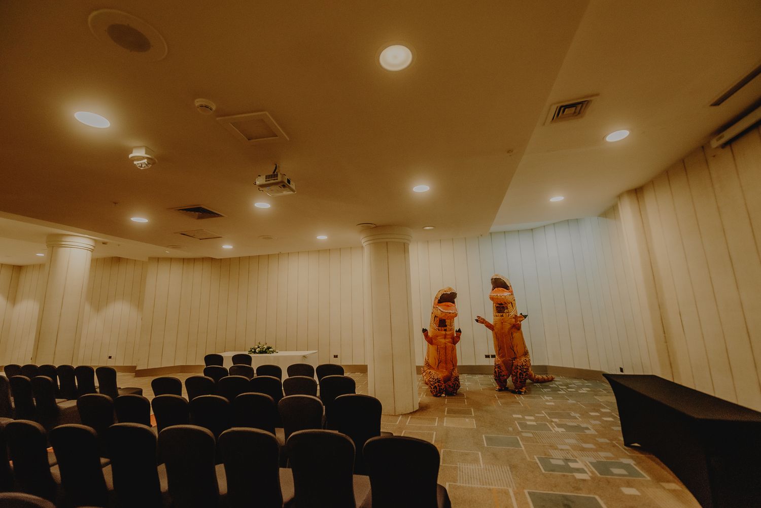 Two people in orange garments stand in an empty auditorium with rows of chairs.