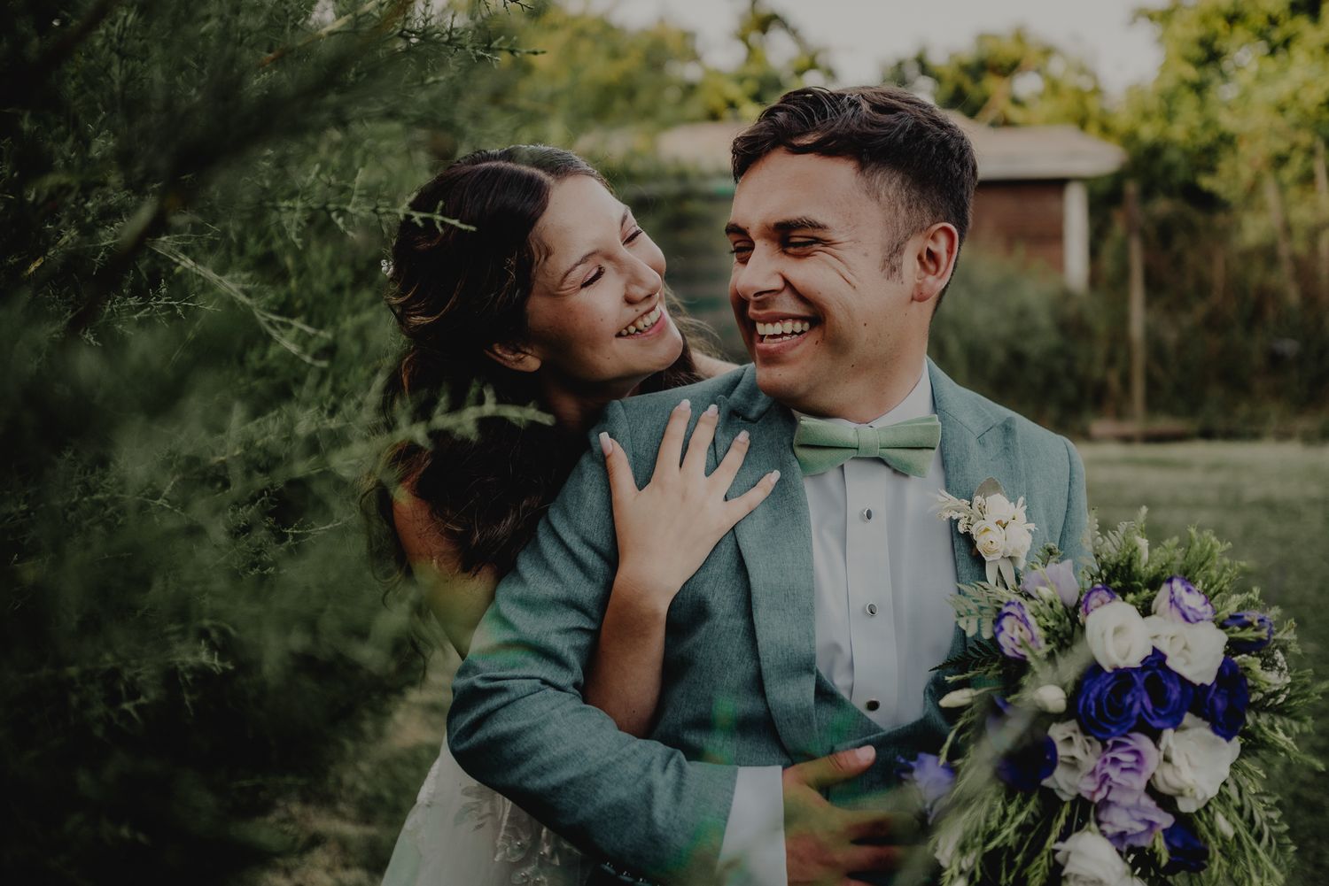 A joyful wedding couple shares a tender moment outdoors while holding purple flowers.