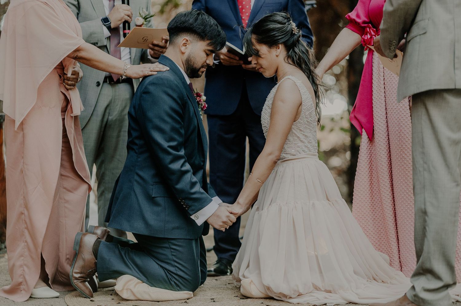 A romantic wedding ceremony moment as a couple kneels together surrounded by guests in soft lighting.