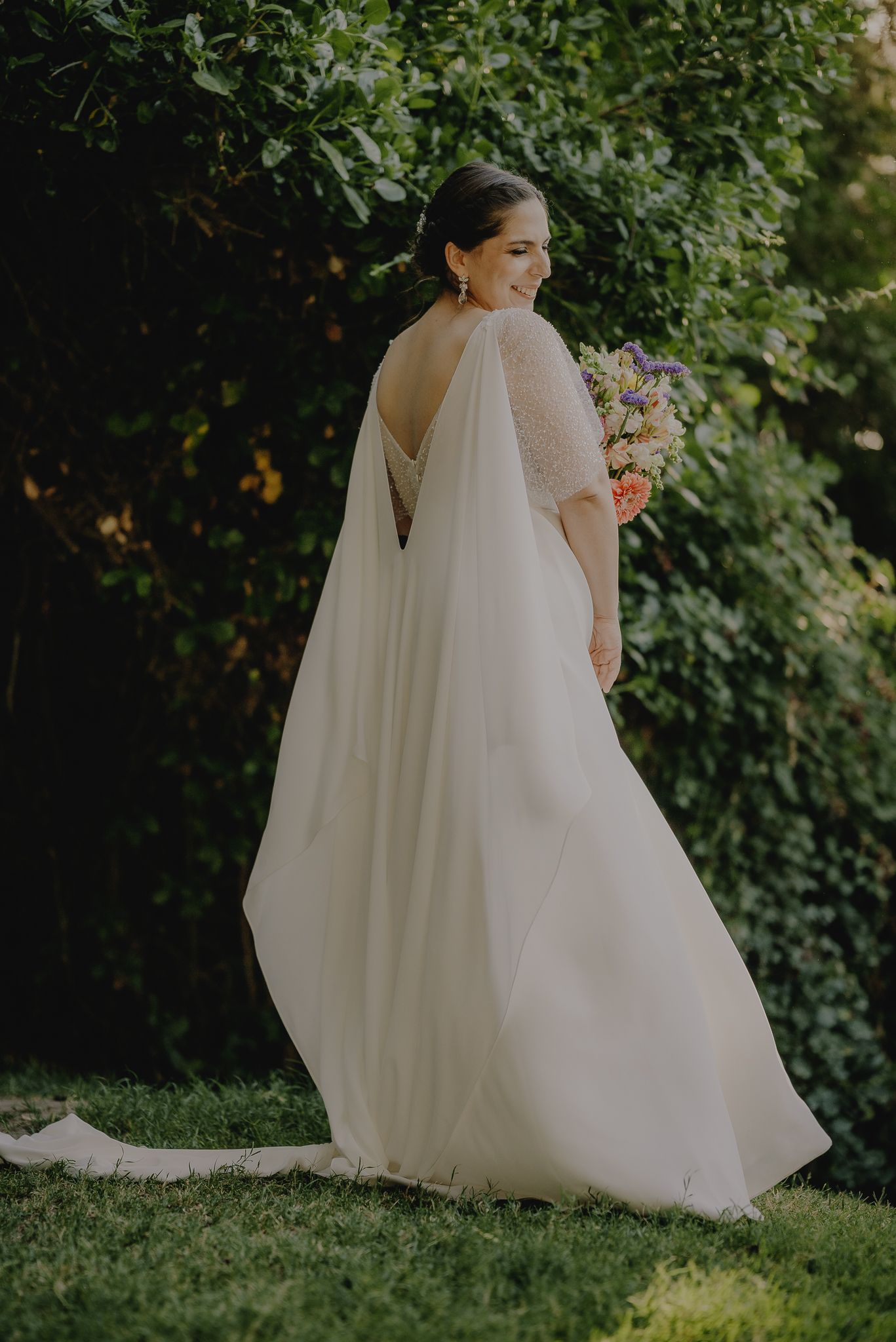 An elegant bride in a flowing white cape dress holds a pastel bouquet in a garden setting.