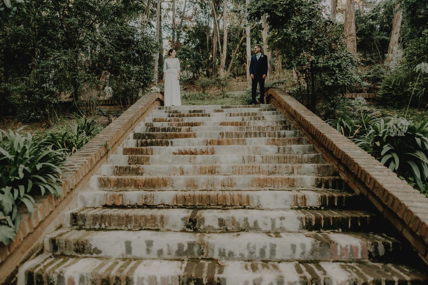 A couple in wedding attire ascends old stone steps surrounded by lush garden foliage.