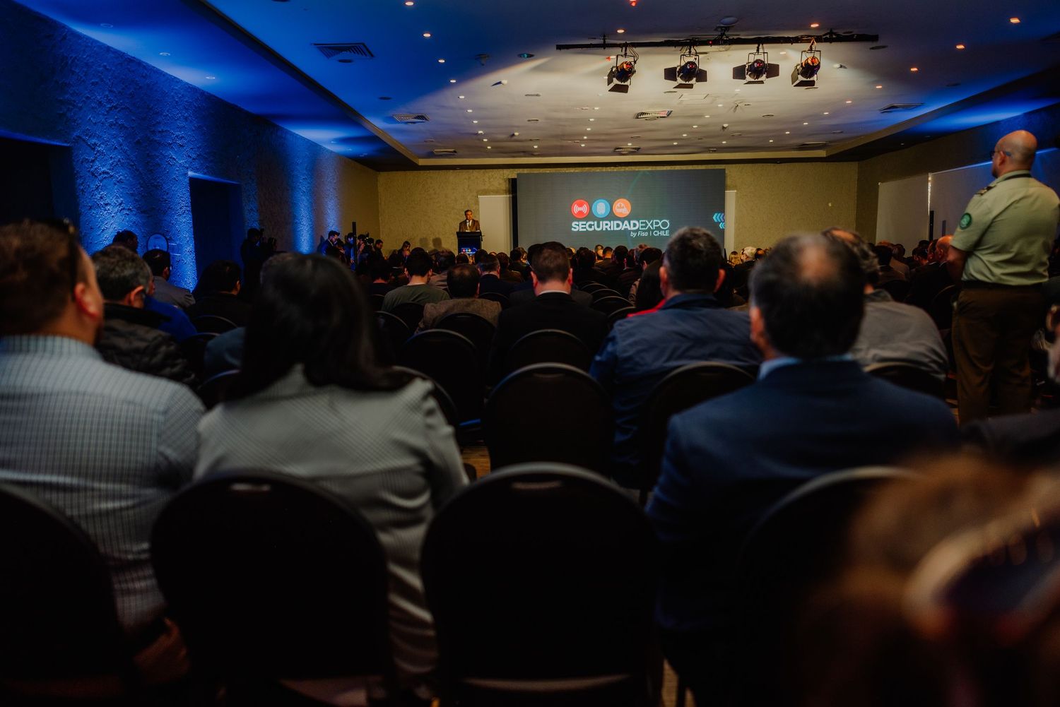 Audience seated at a professional conference event with blue lighting and a presentation screen at the front of the room.