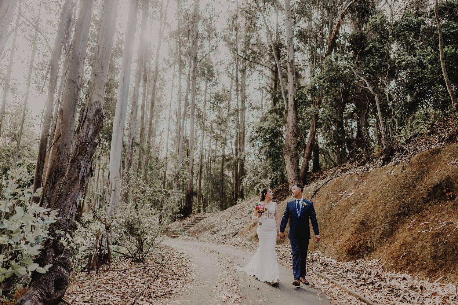 A couple in wedding attire walks hand in hand along a forest path surrounded by tall eucalyptus trees.