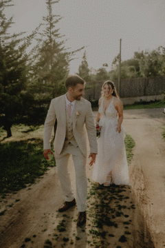 Couple walking hand in hand on sunlit path with bride in flowing white dress.