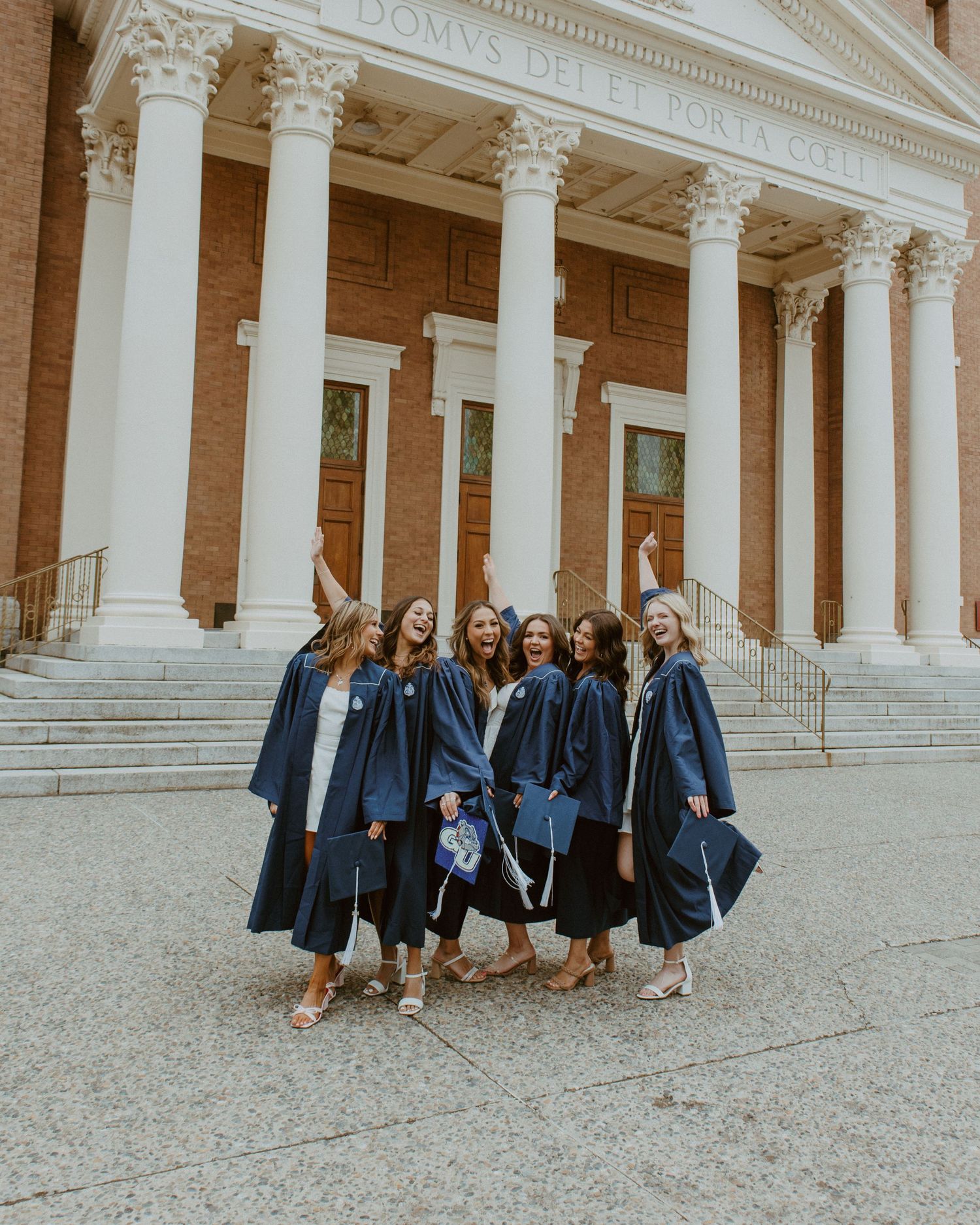 A group in graduation caps and gowns poses on the steps of a grand building with white classical columns in warm light.