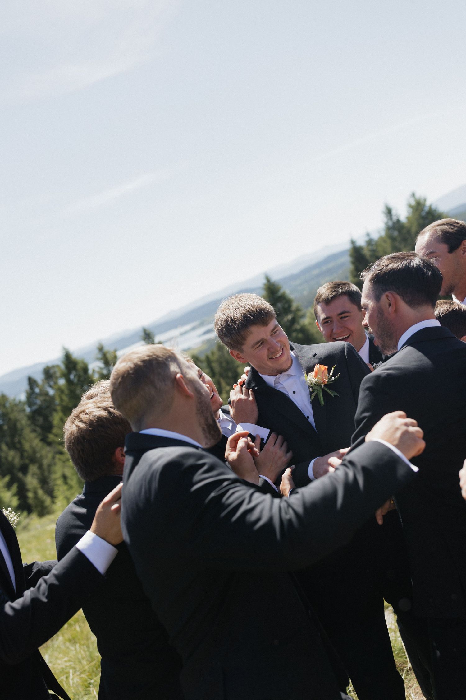 A group of groomsmen in black suits take shots of alcohol together at an outdoor wedding with mountains in background.
