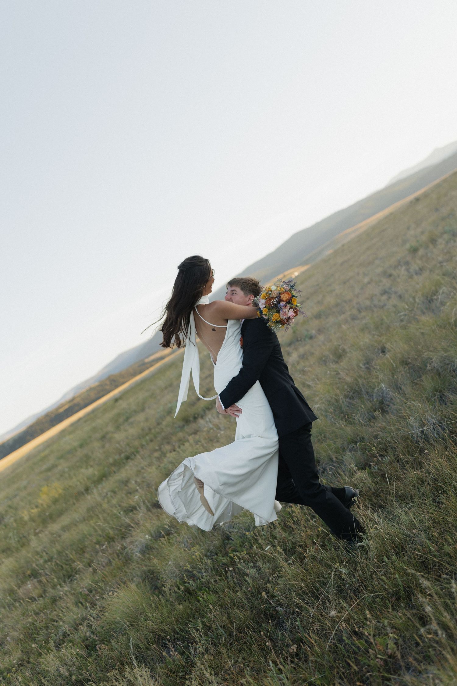 Two figures embrace while sitting on a grassy hillside during golden hour.