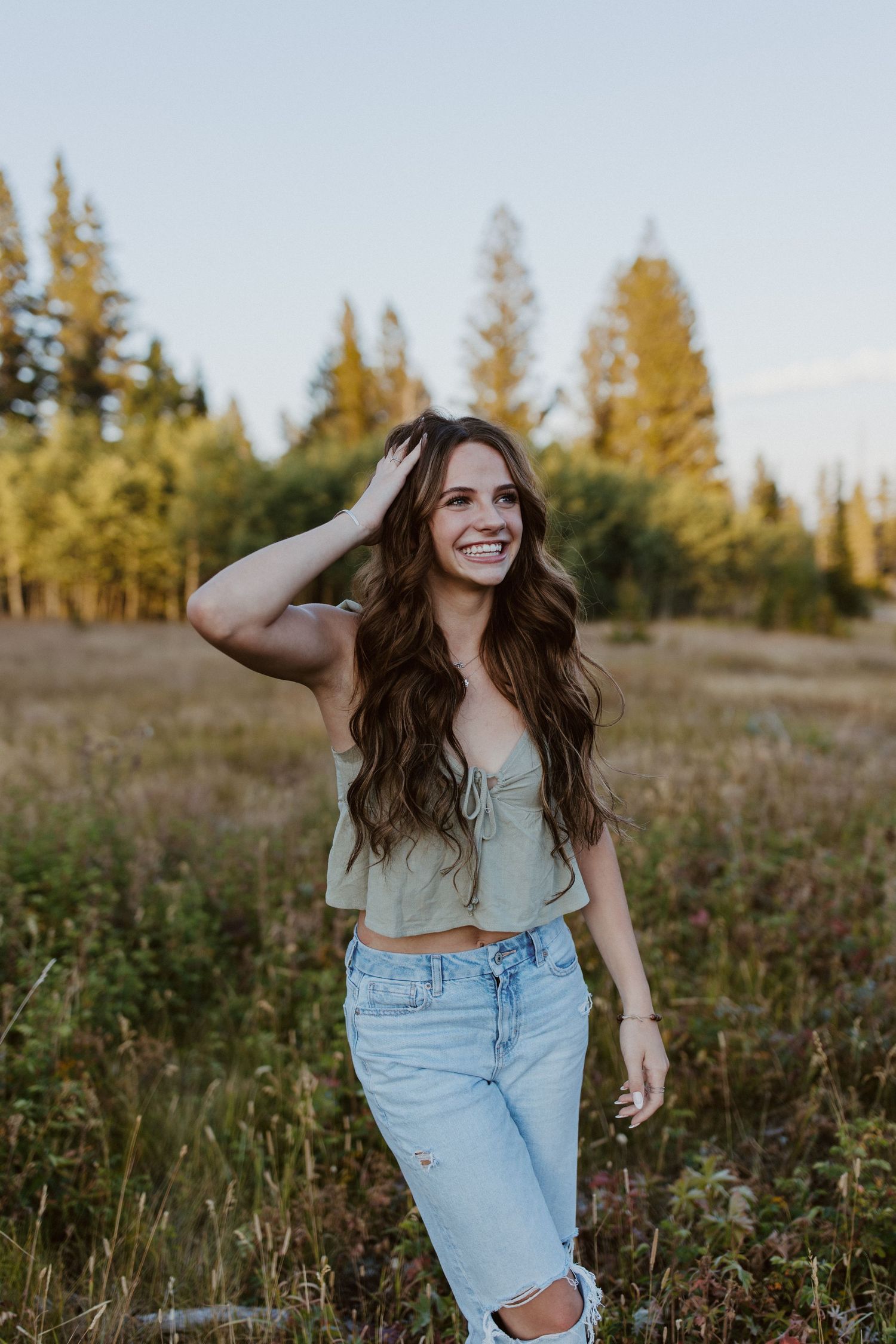 Woman in crop top and ripped jeans walks through a golden field at sunset with evergreen trees in background.