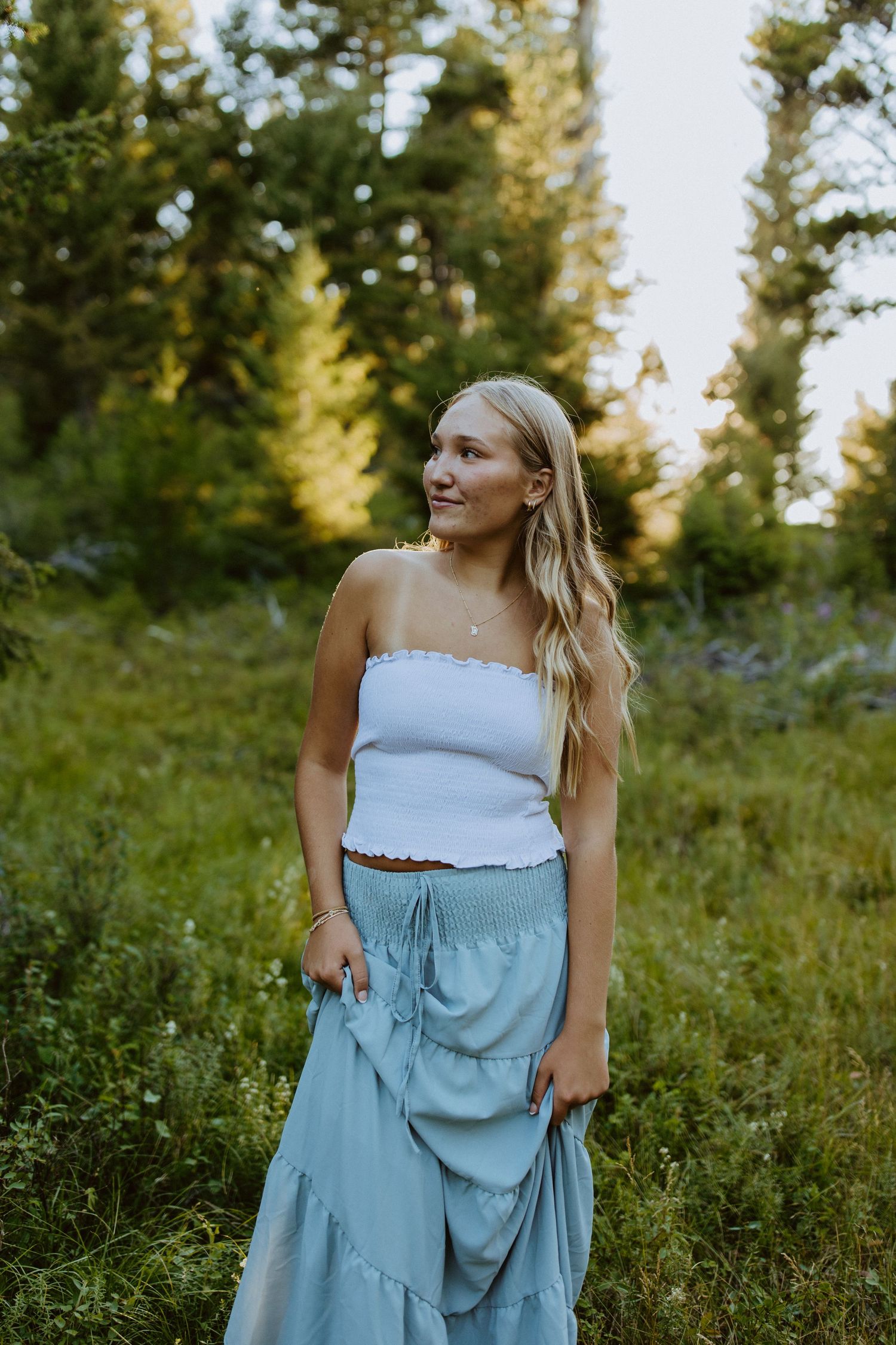 A woman in a white tube top and light blue maxi skirt smiles while standing in a sunlit meadow with evergreen trees.