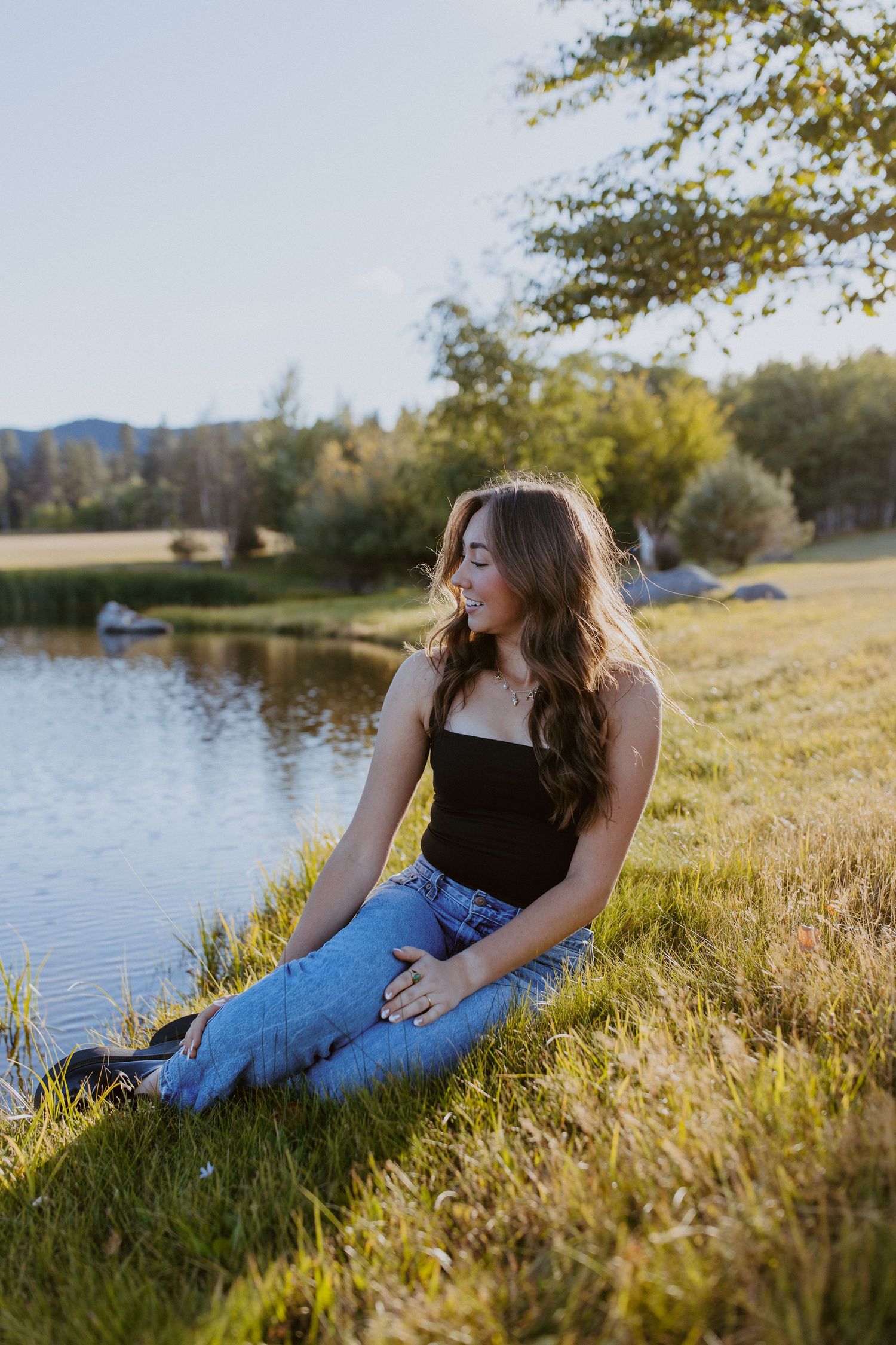 Person sitting in casual attire by a serene lake at sunset with mountains and trees in the background.