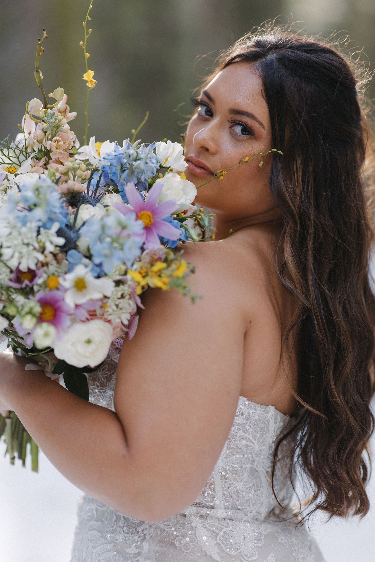 A bride in a white lace dress holds a vibrant bouquet with blue and white flowers against a soft blurred background.