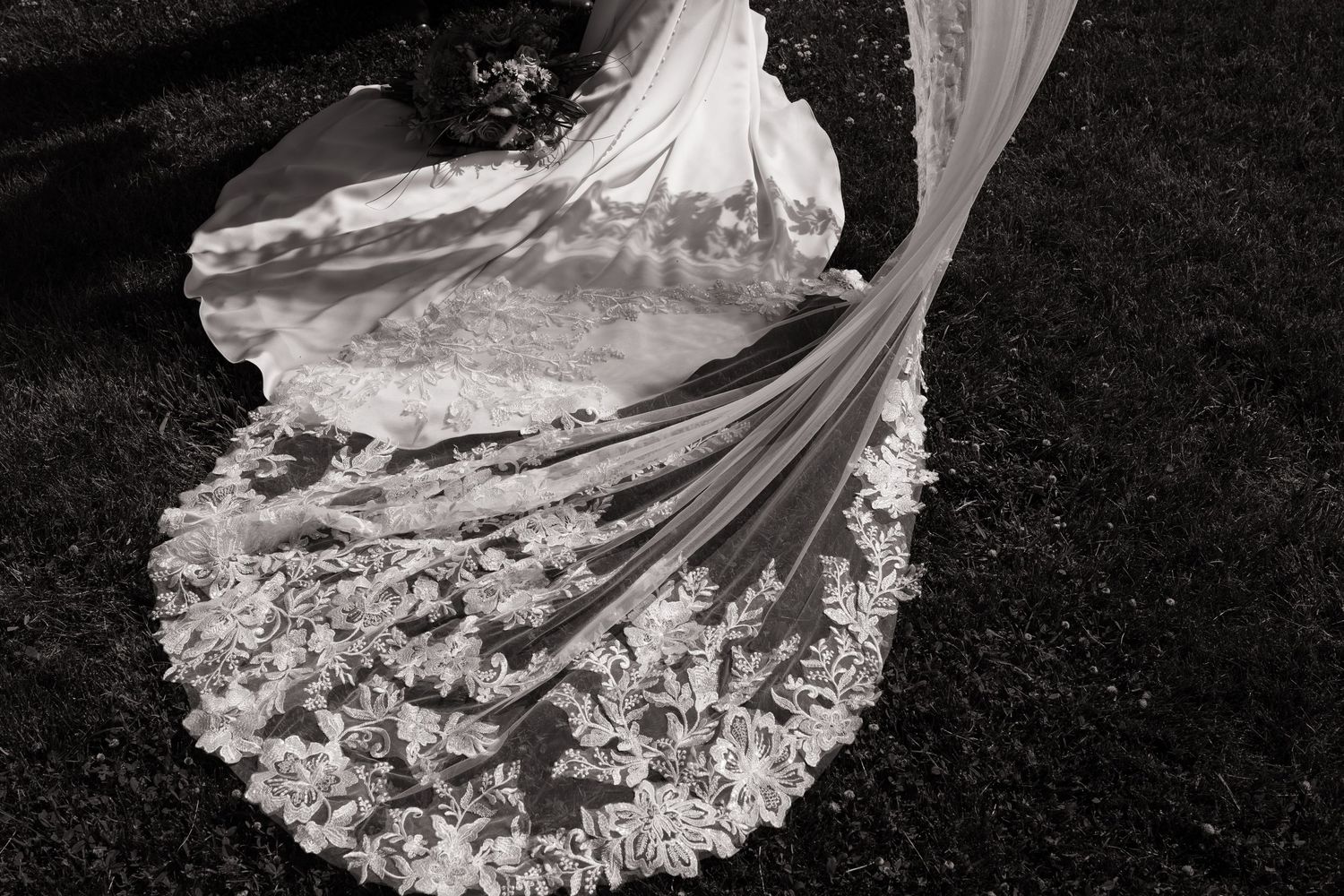 An elegant bridal gown with intricate lace trim and flowing train photographed in black and white on grass.