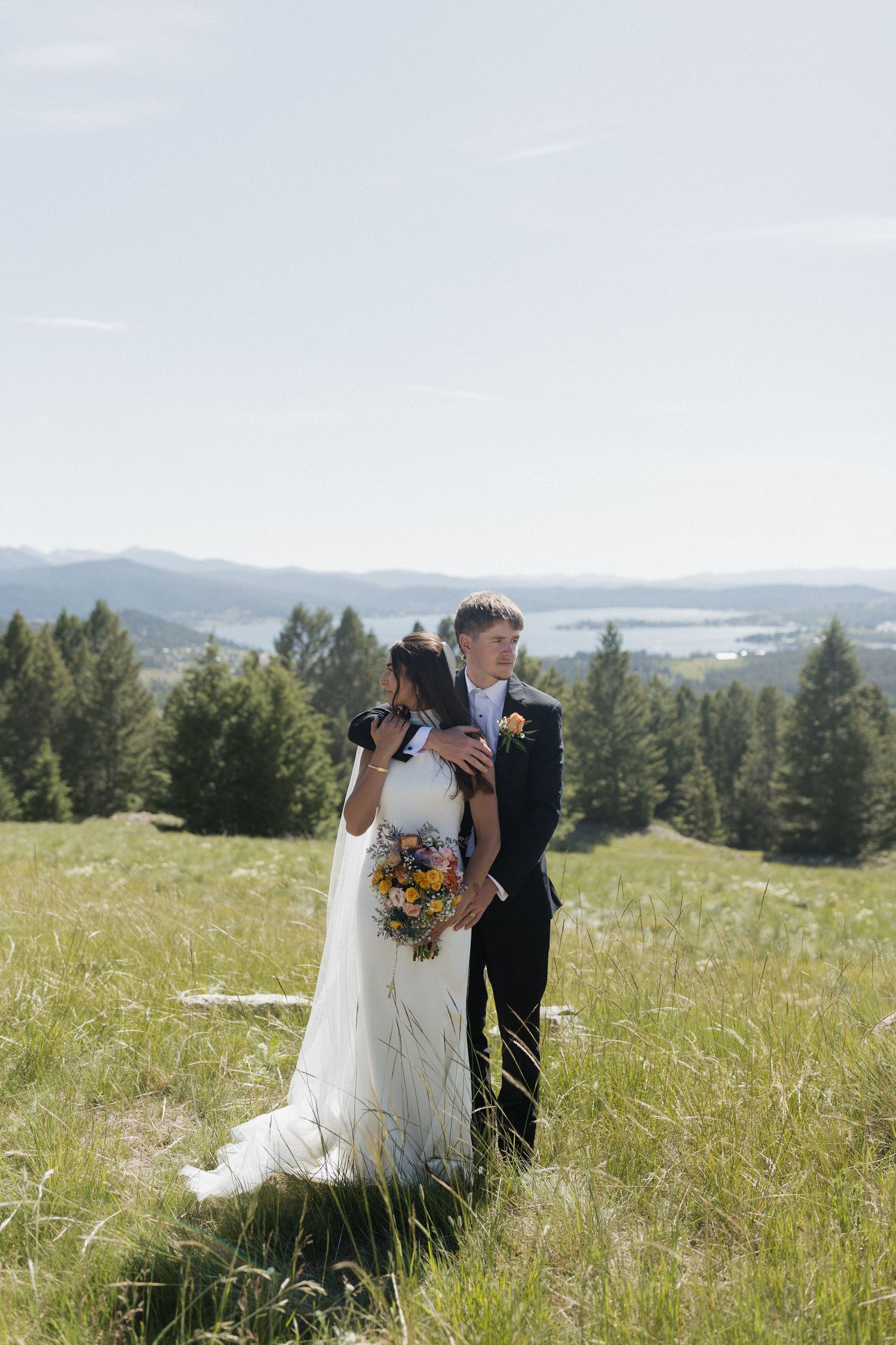 A couple celebrates their wedding day in a scenic mountain meadow overlooking a lake and evergreen forest.