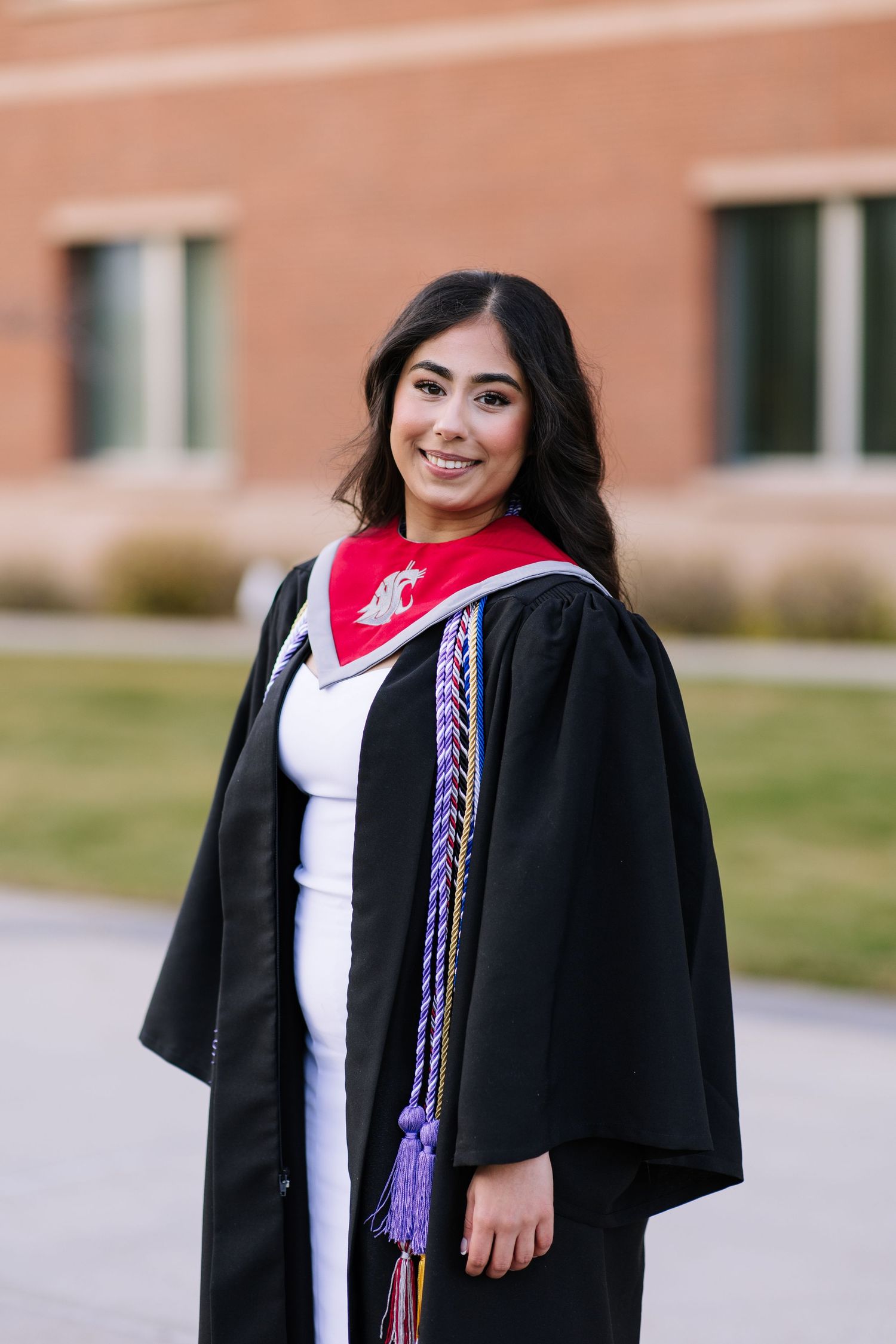 Graduate wearing a black academic gown with red stole stands outdoors by a brick building on graduation day.