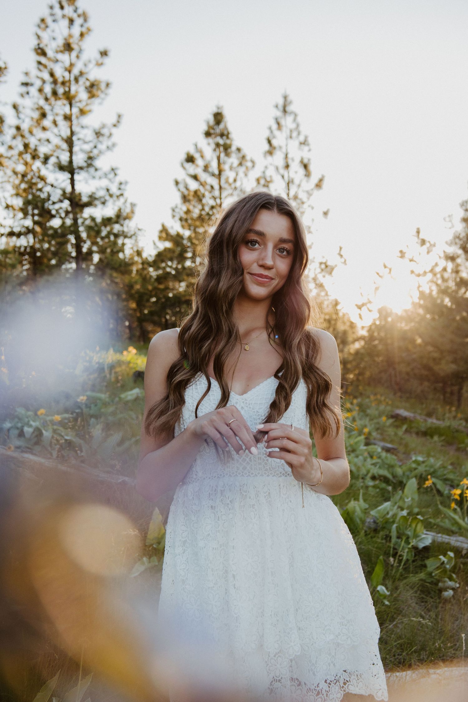 A delicate white lace dress glows in golden sunset light among pine trees and wildflowers in a natural outdoor setting.