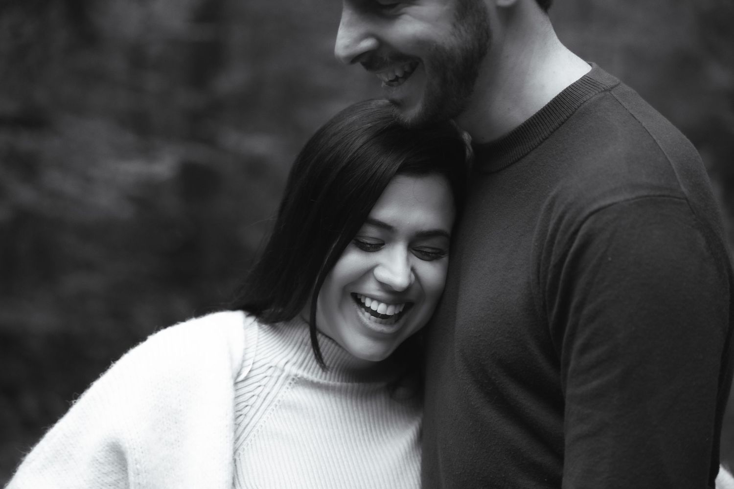 Black and white photo of a laughing couple embracing outdoors, woman in white sweater leaning on smiling man.