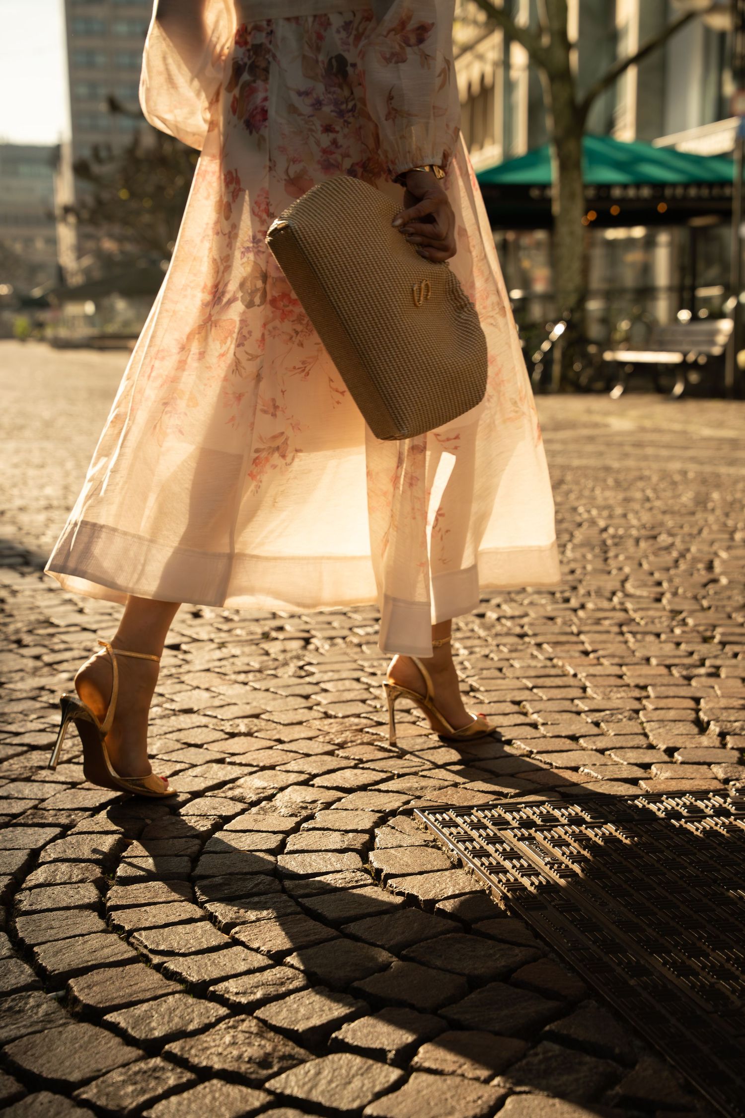 Woman in floral dress and heels carrying a woven clutch bag on cobblestone street in golden sunlight.
