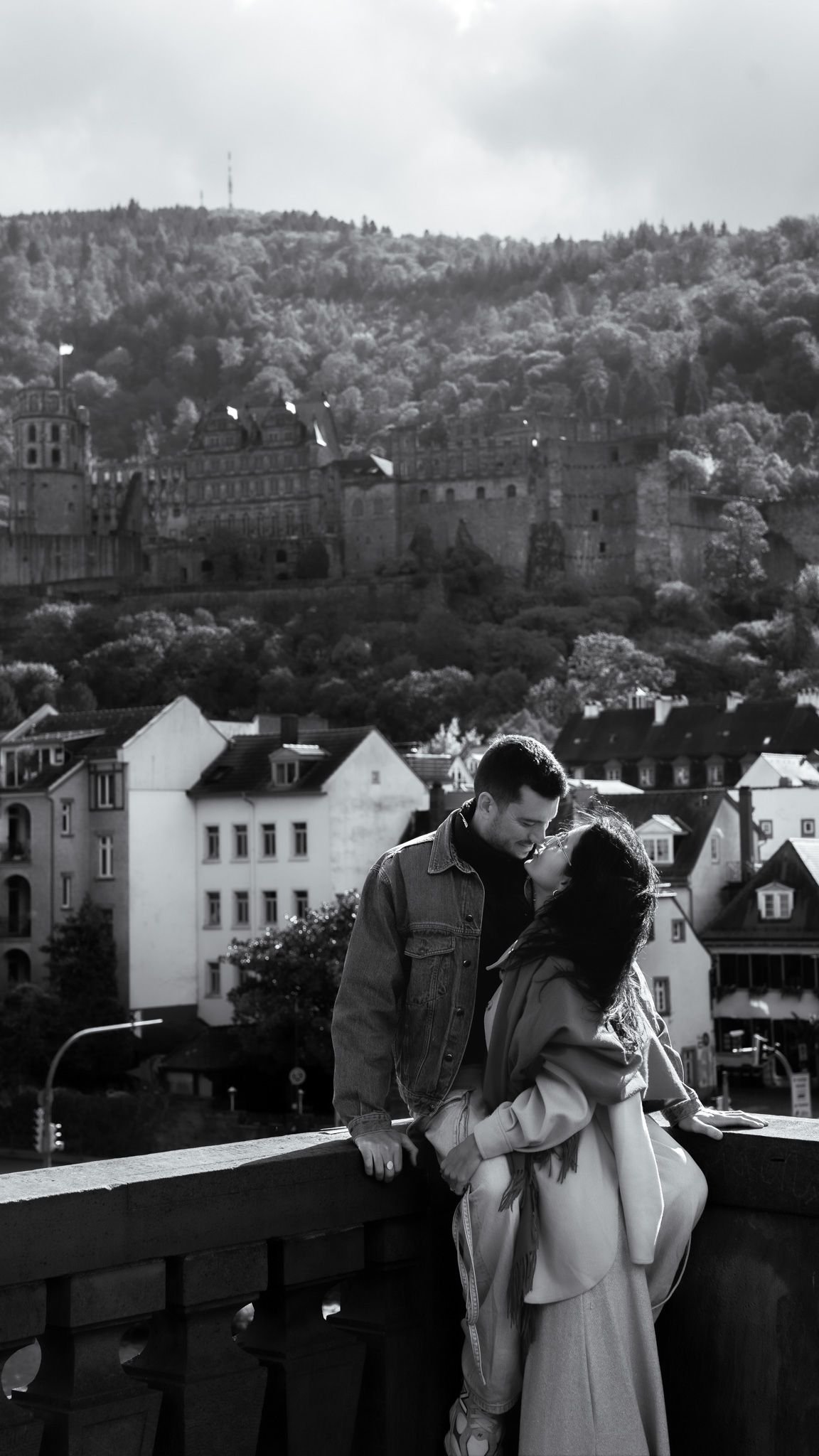 A romantic couple embraces on a balcony overlooking a European cityscape with historic buildings and hillside views.