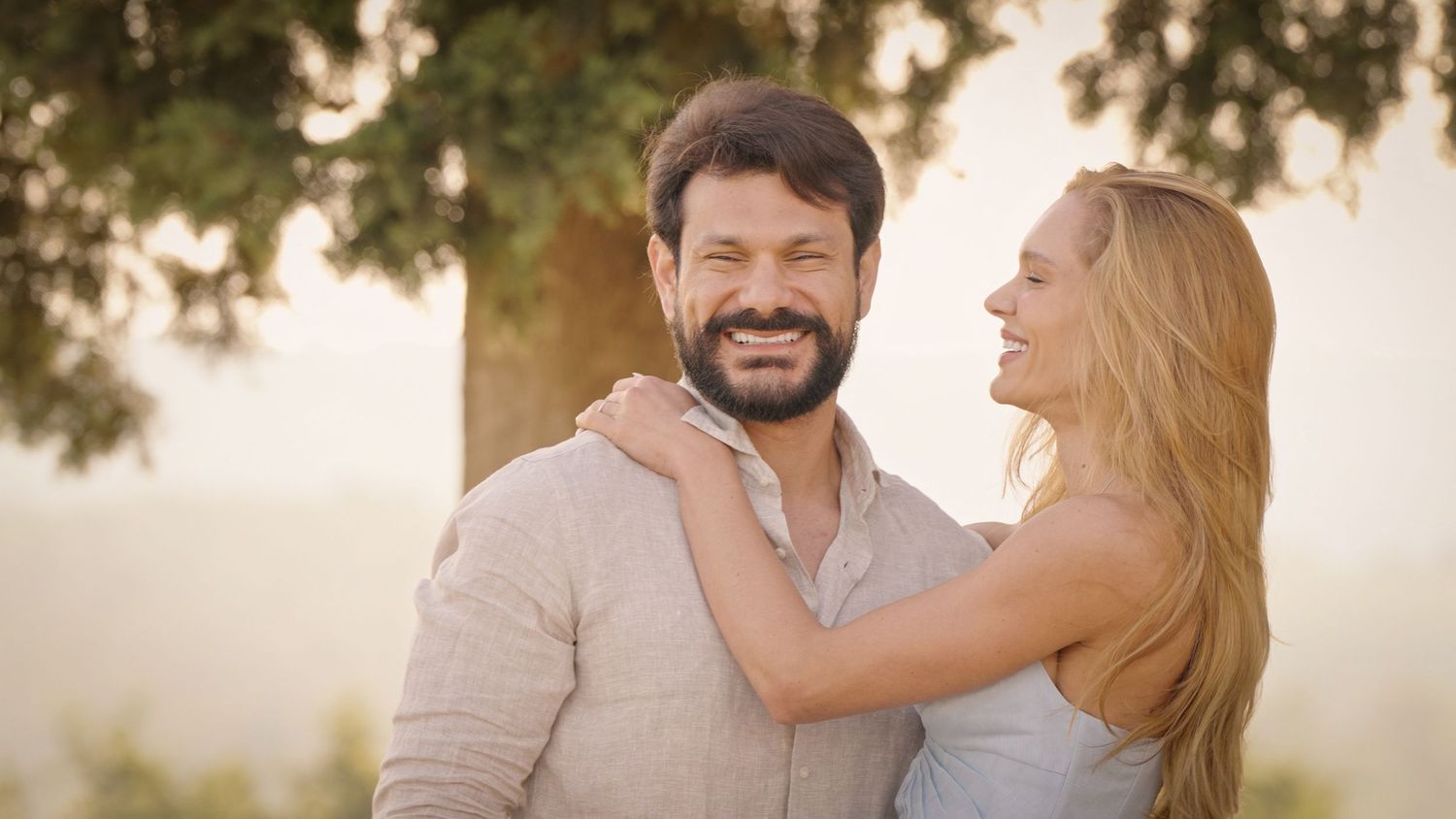 Happy couple embracing outdoors under a tree in warm golden sunlight, smiling man with beard and blonde woman.