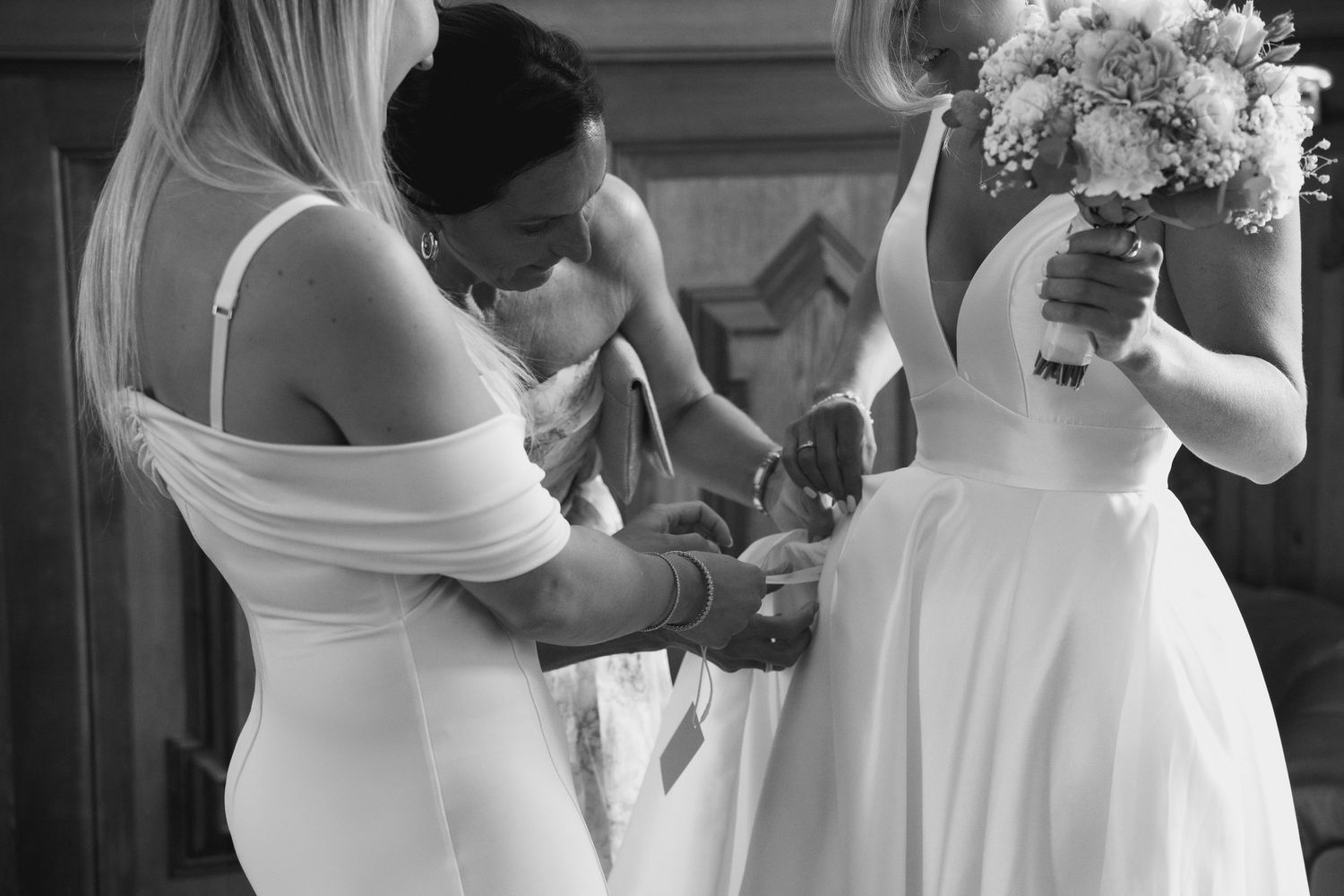 Bridesmaids helping bride with dress details while holding bouquet, black and white wedding preparation photo.