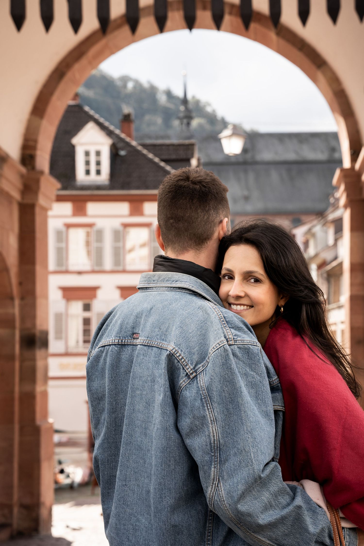 A couple in a romantic embrace stands beneath stone archways in a European town setting.