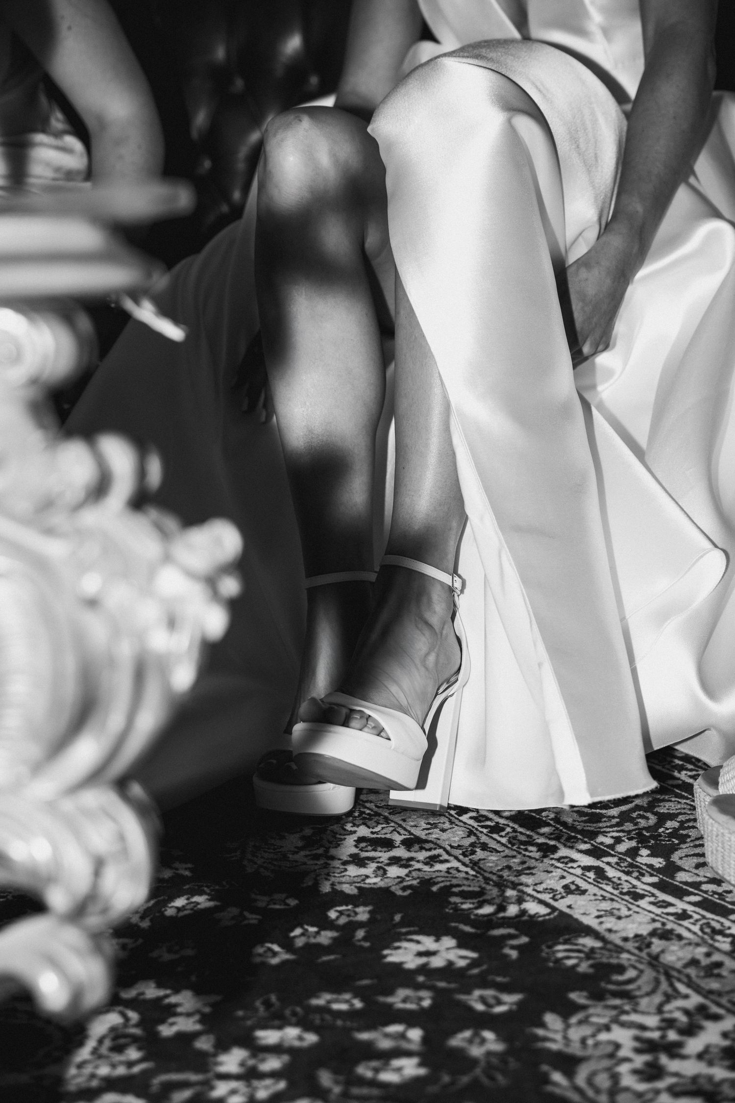 Black and white photo of a bride in white dress and sandals seated near ornate furniture on a patterned rug.