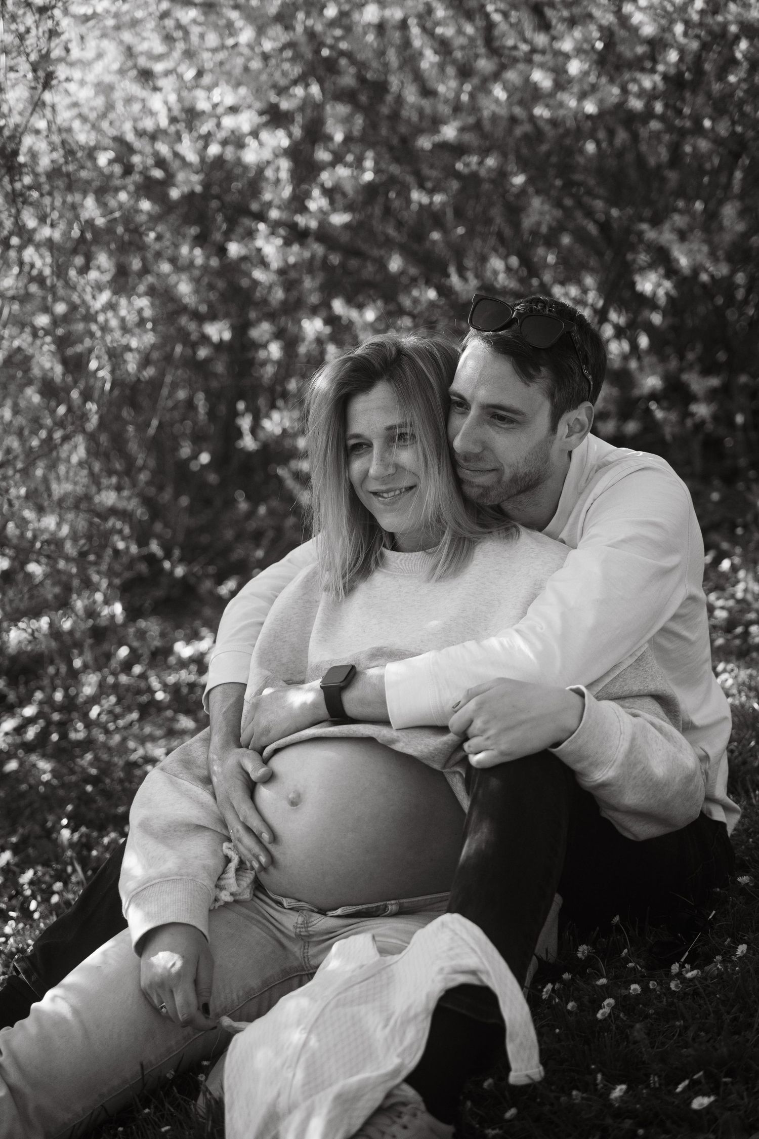 Black and white maternity photo of a couple embracing outdoors with blooming trees in the background.