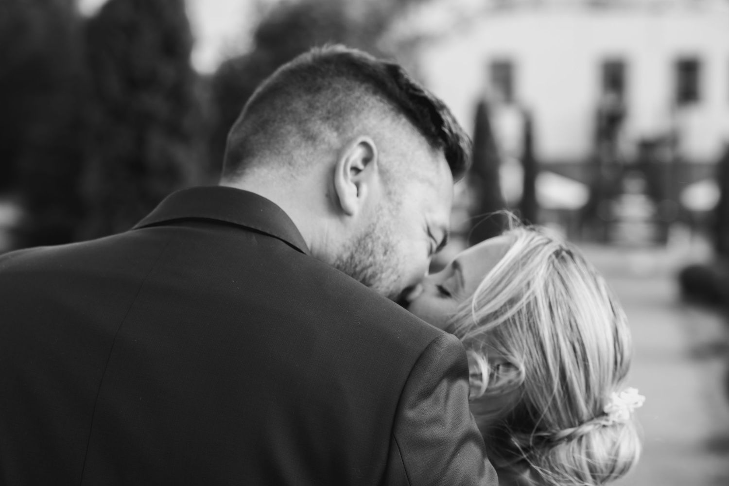 Black and white photo of a couple sharing a romantic kiss, bride with updo hairstyle and groom in suit.