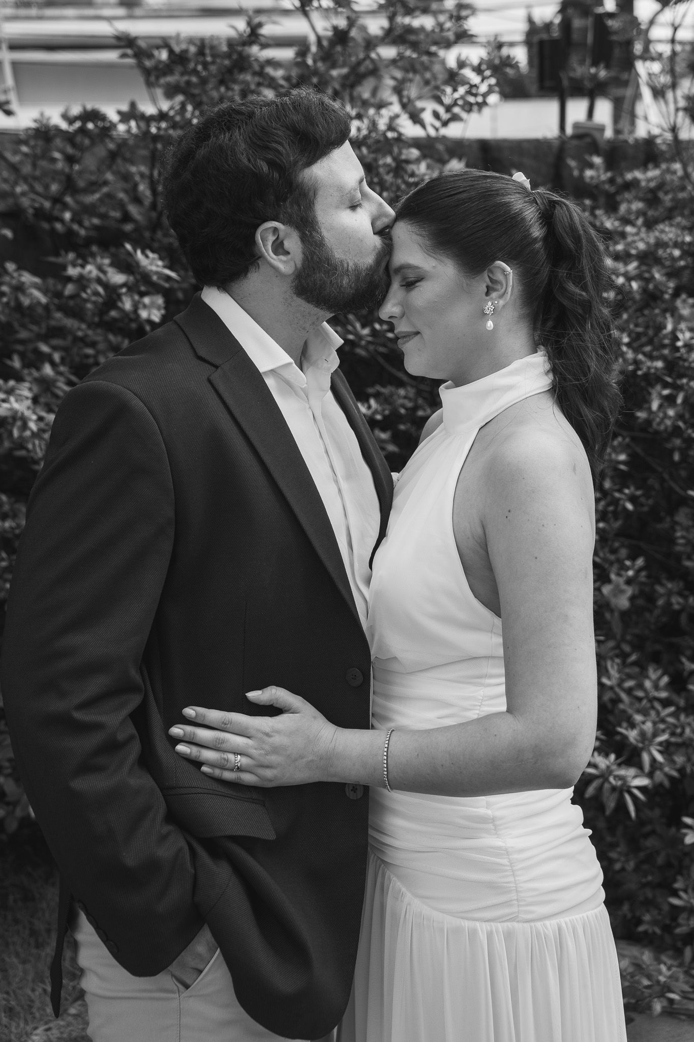 Black and white photo of a couple in wedding attire sharing a romantic moment outdoors near lush greenery.