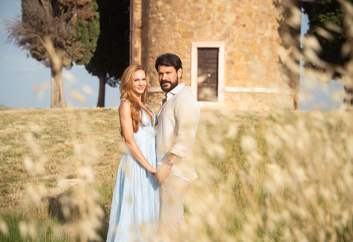 A couple embraces in a sun-drenched field beside a rustic stone building on a warm summer day.