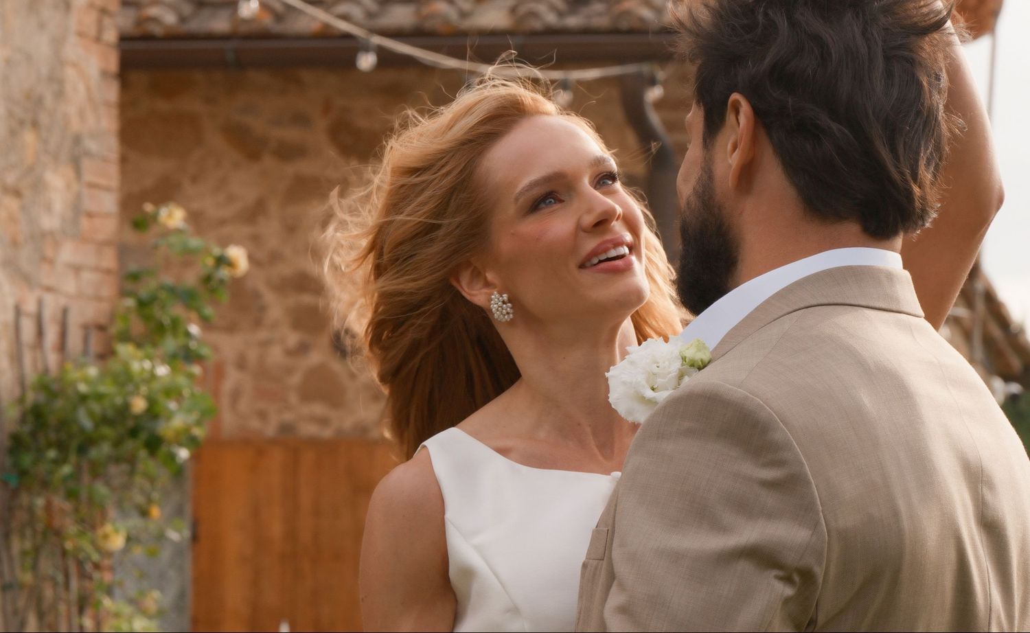 Bride in white dress smiles at groom in tan suit during outdoor wedding ceremony.