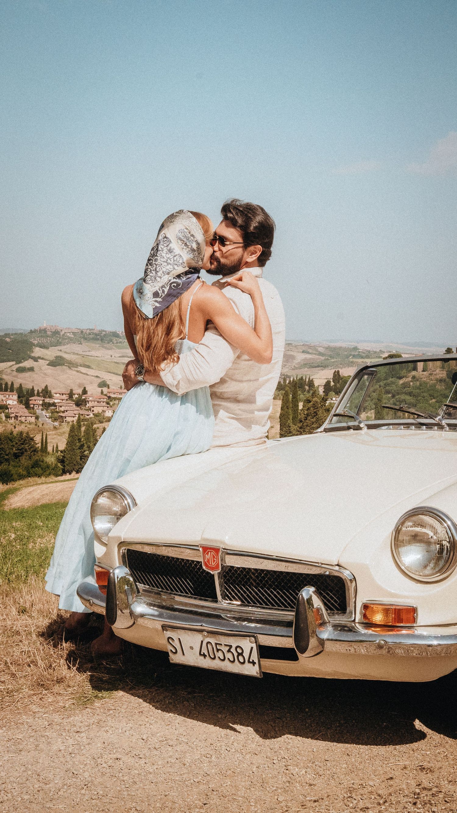 Couple embracing romantically on a vintage white MG convertible in the Tuscan countryside on a sunny day.