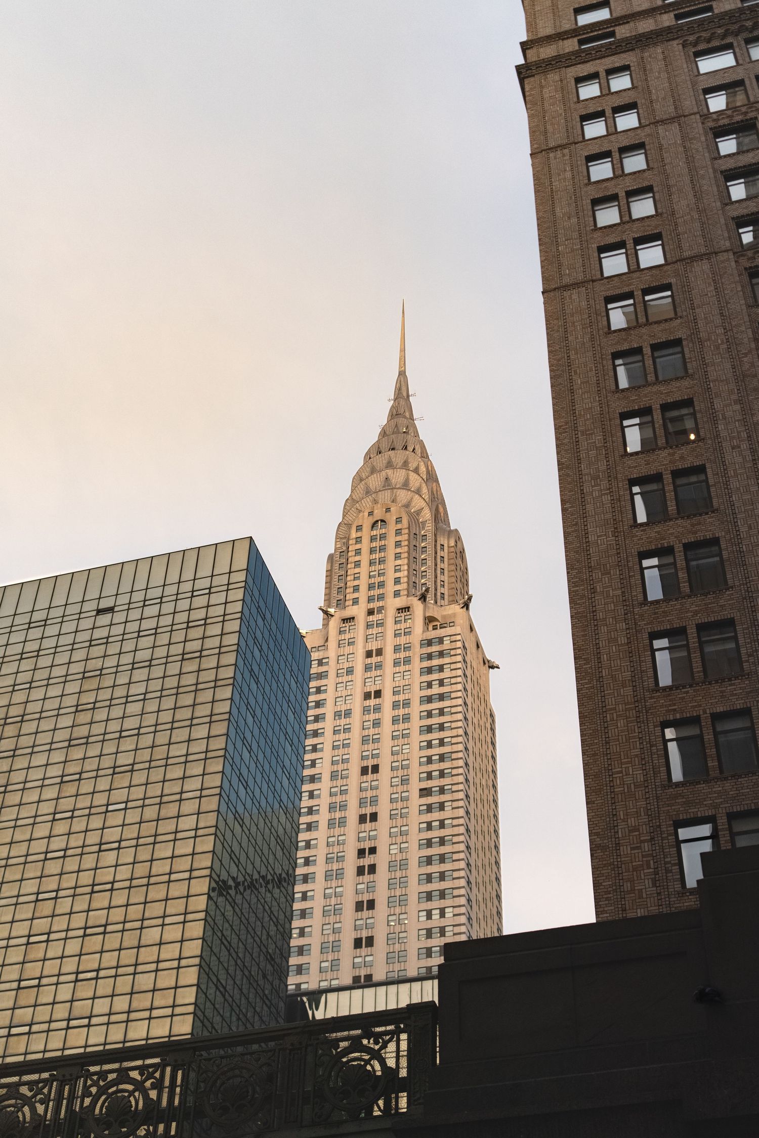 Iconic Chrysler Building spire and modern glass buildings in New York City against cloudy sky.