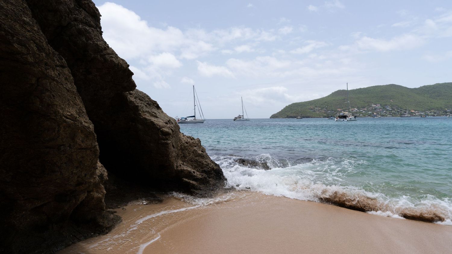 Rocky coastal cove with waves crashing against cliffs and boats visible in the distance.