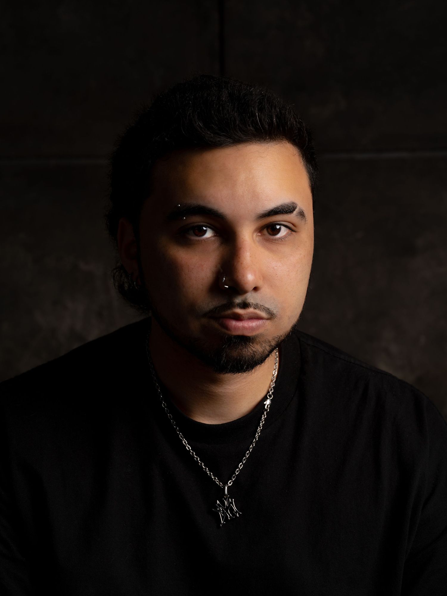 Young man with nose ring and chain necklace wearing black shirt poses against dark studio background.