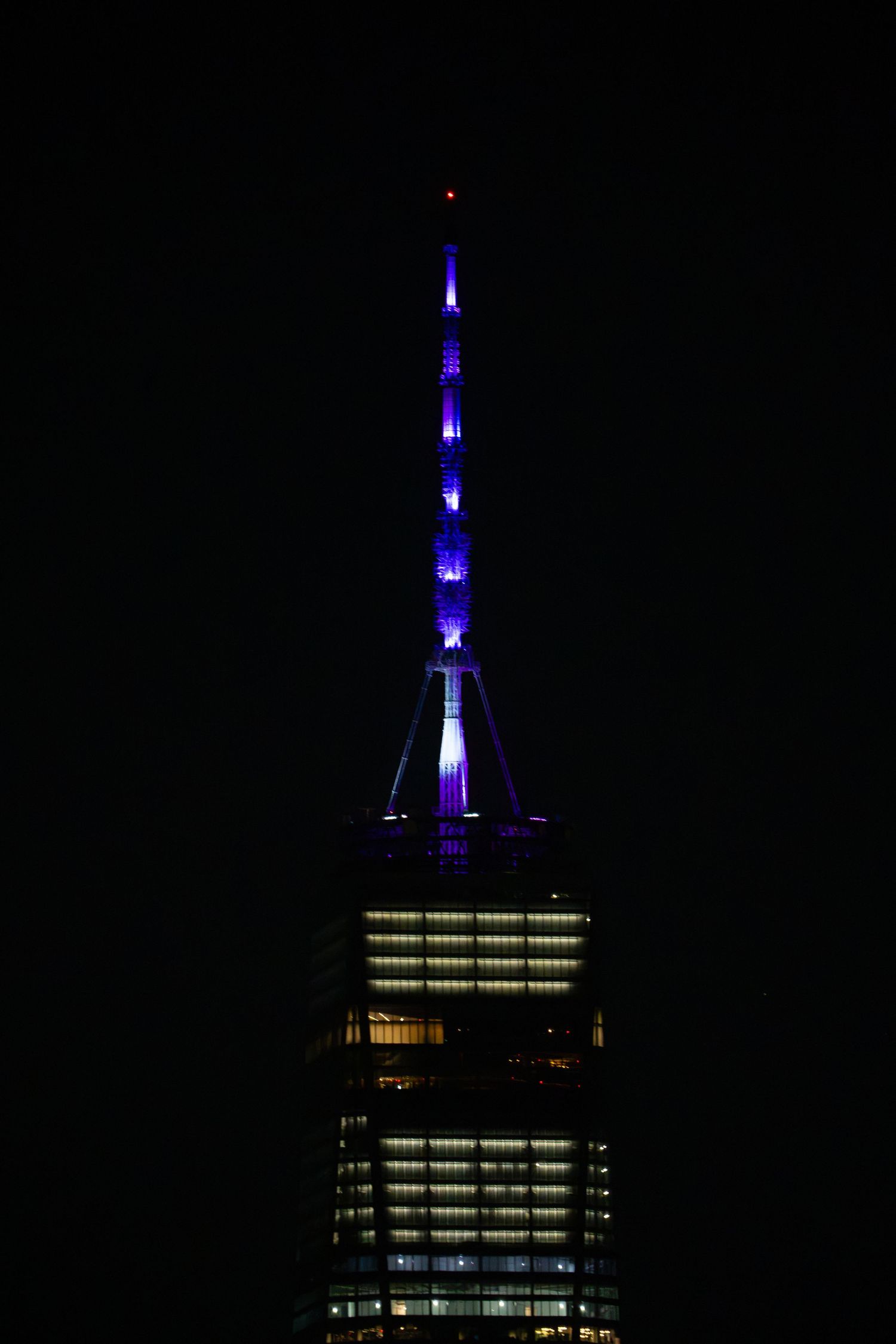 Empire State Building spire illuminated in purple and blue lights against dark night sky.
