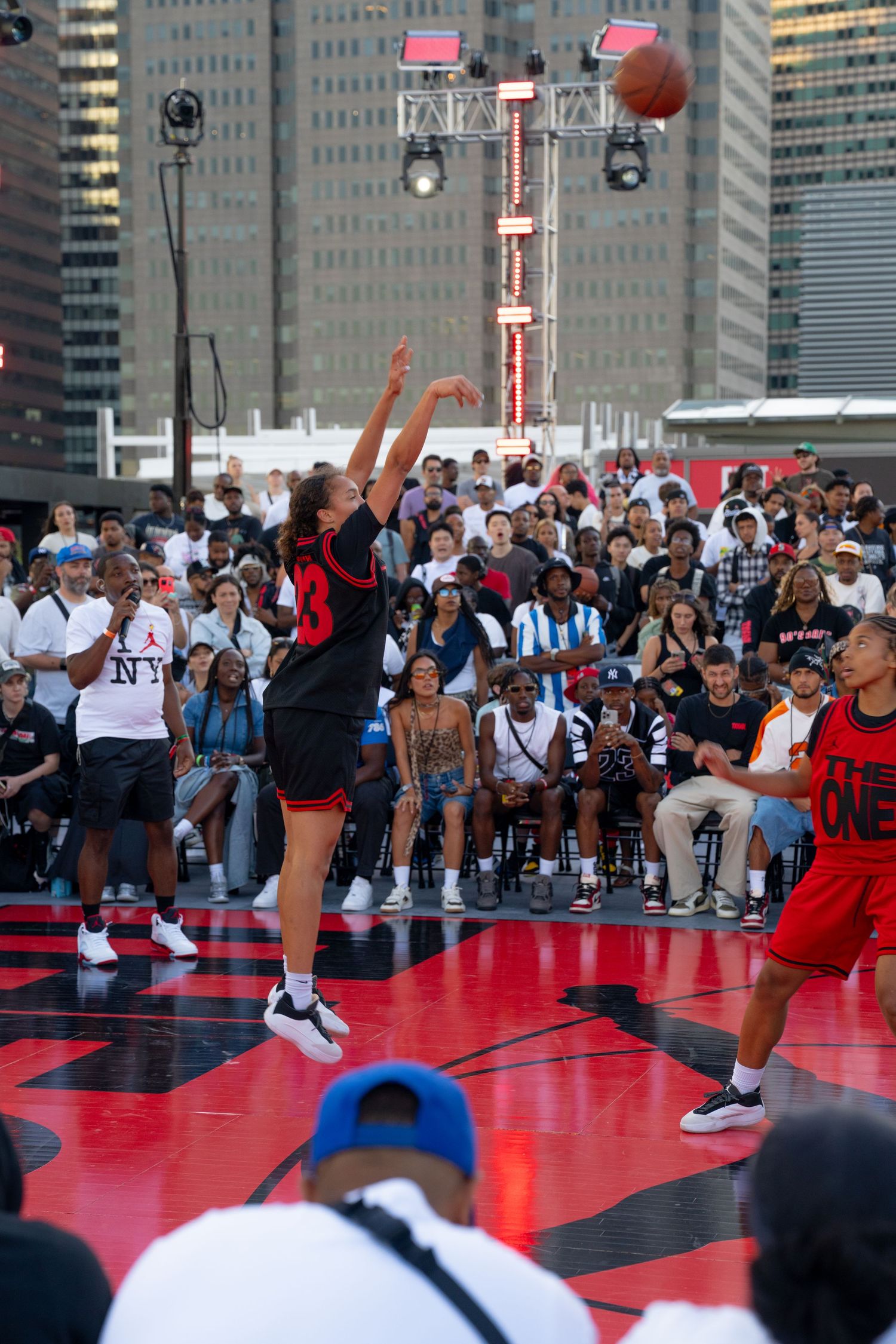 Basketball players competing on a red outdoor court with city skyscrapers in the background.