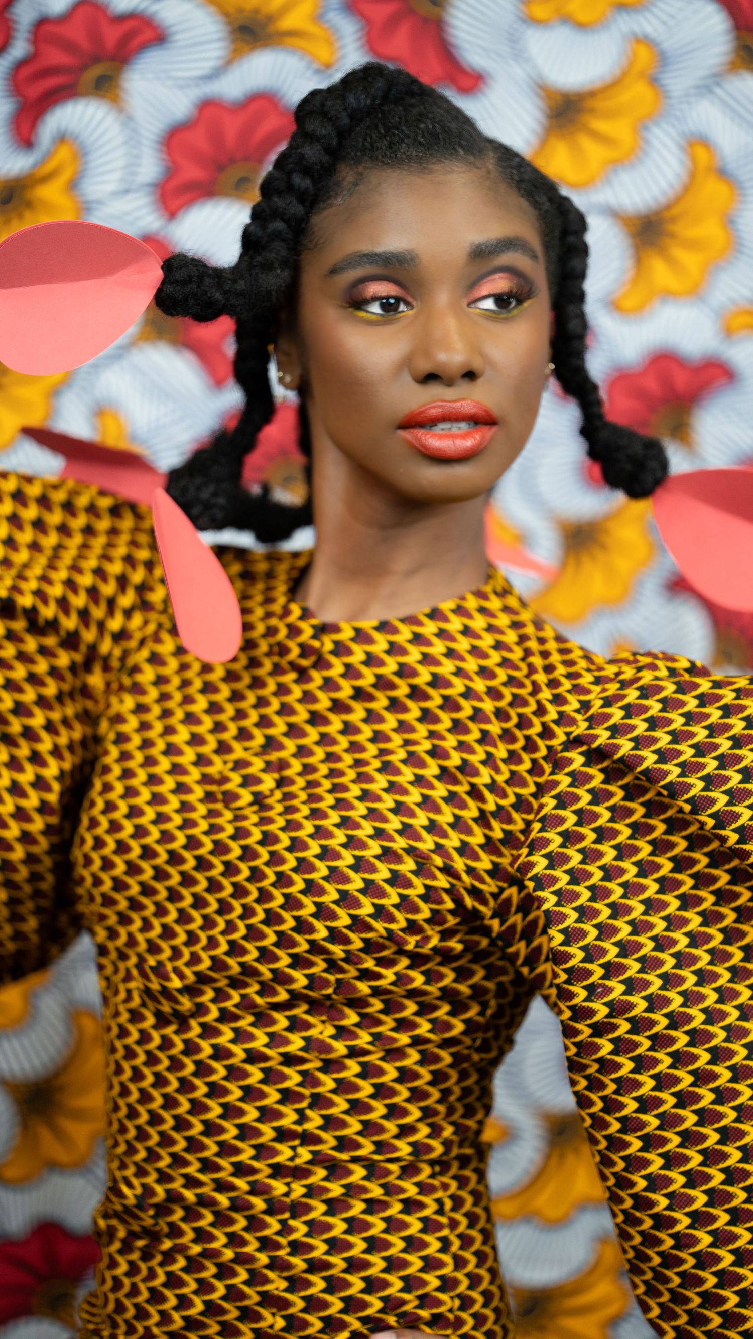 Model in yellow patterned dress poses against floral backdrop with vibrant makeup.