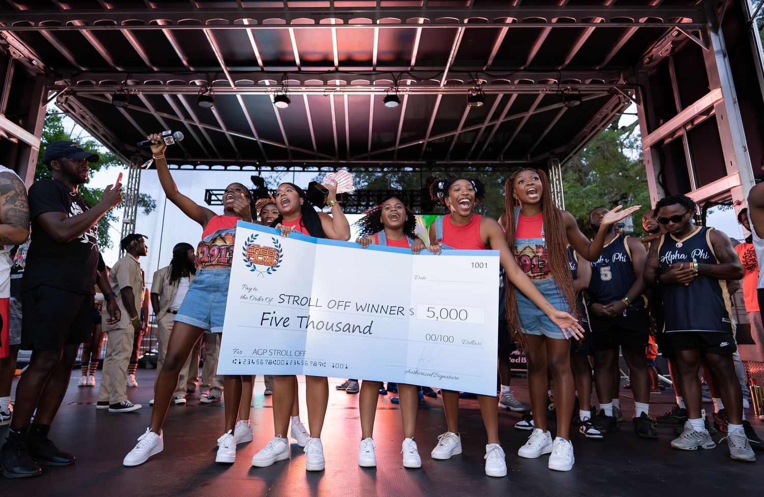 Large group of people gathered under a covered outdoor venue holding oversized checks and celebrating.