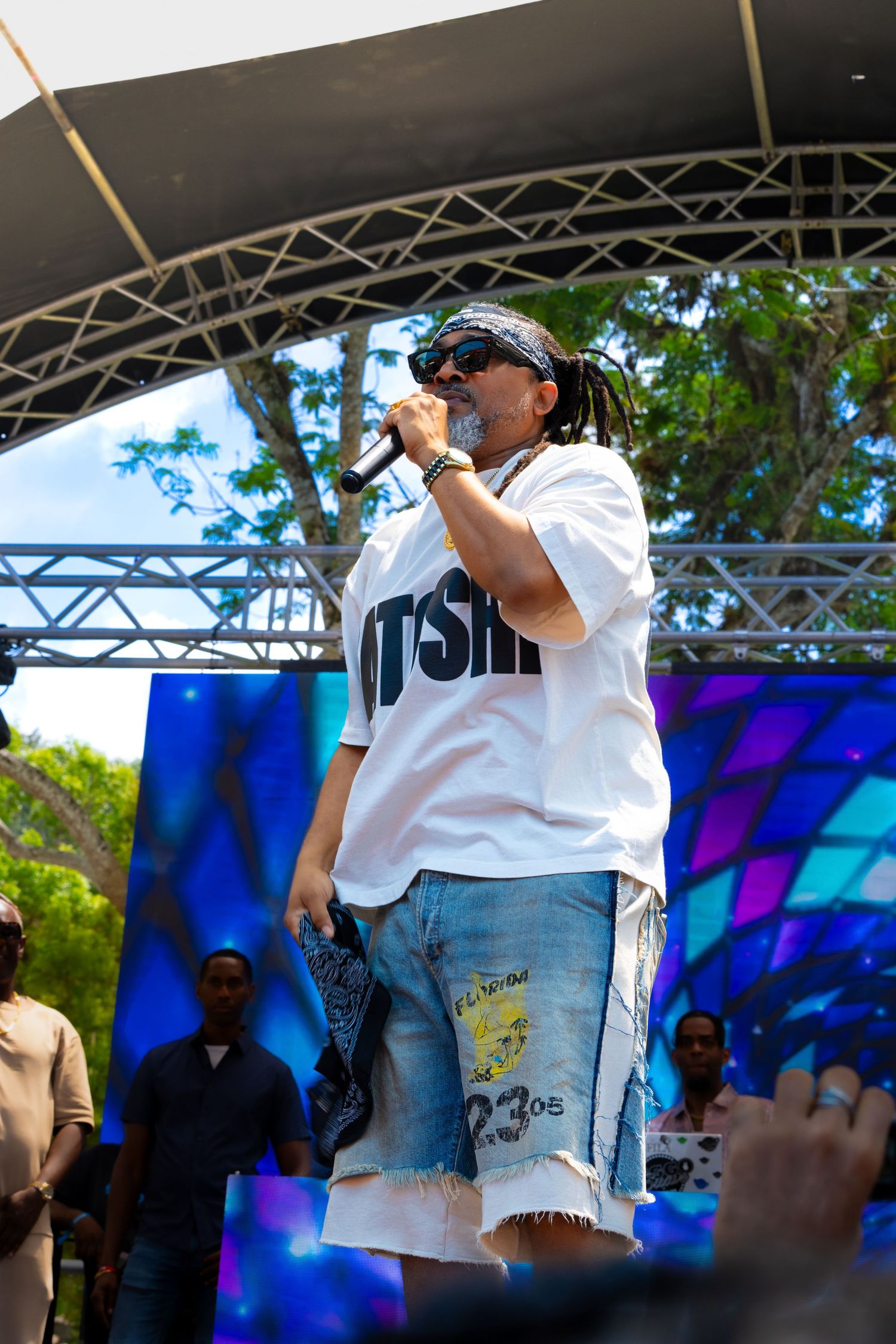 Performer in white shirt and sunglasses singing on outdoor stage with colorful backdrop.