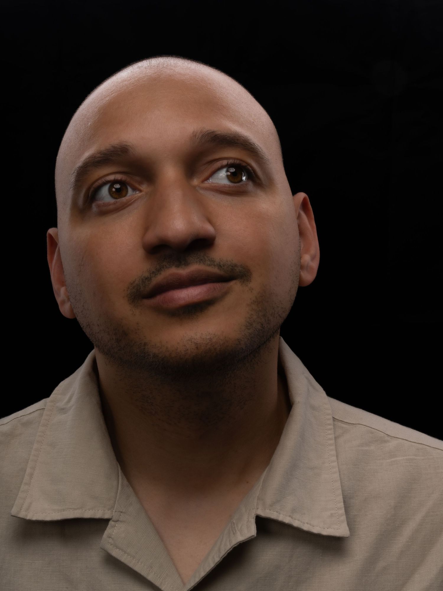 Bald man with goatee looking upward against dark background, wearing beige collared shirt.
