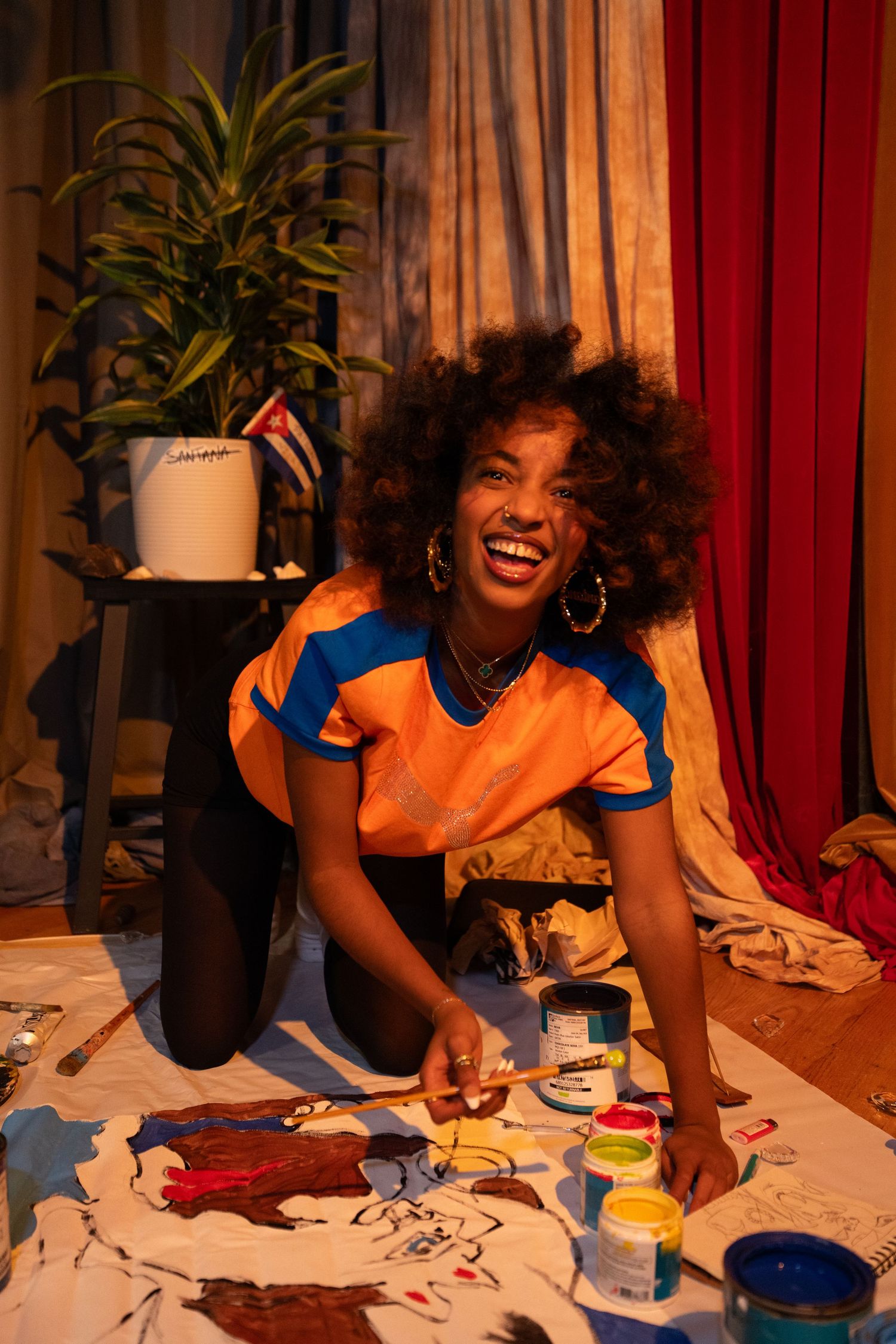 Artist wearing orange and blue outfit sits on floor with paint supplies and smiles while creating colorful artwork.