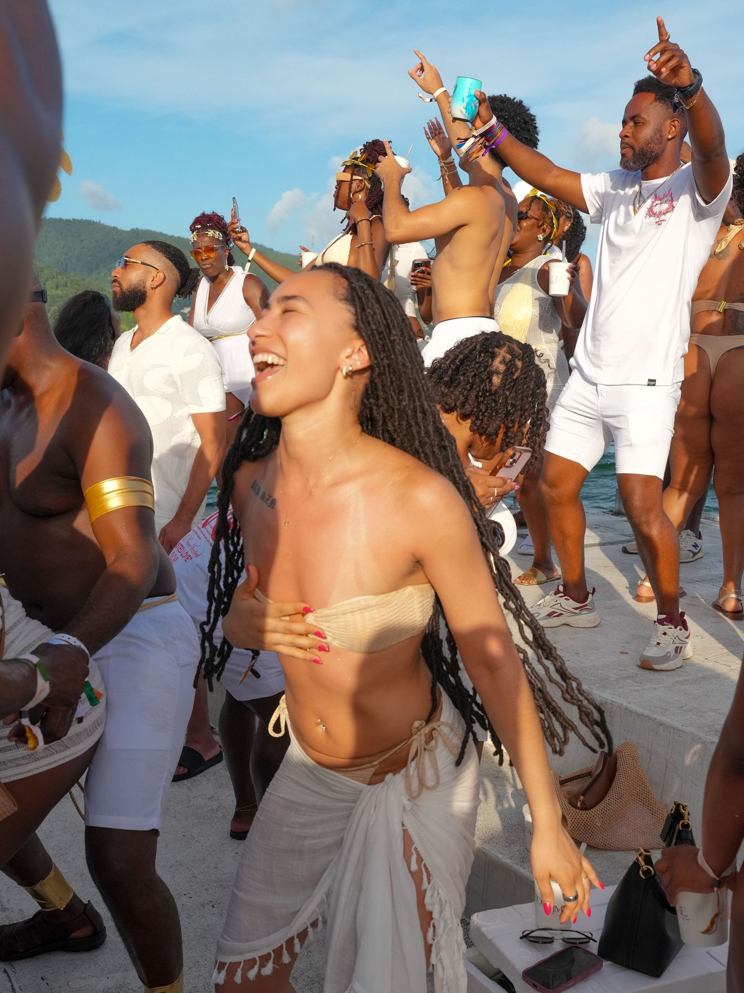 Group of people dancing and celebrating outdoors at a beach party while holding drinks in the sunlight.
