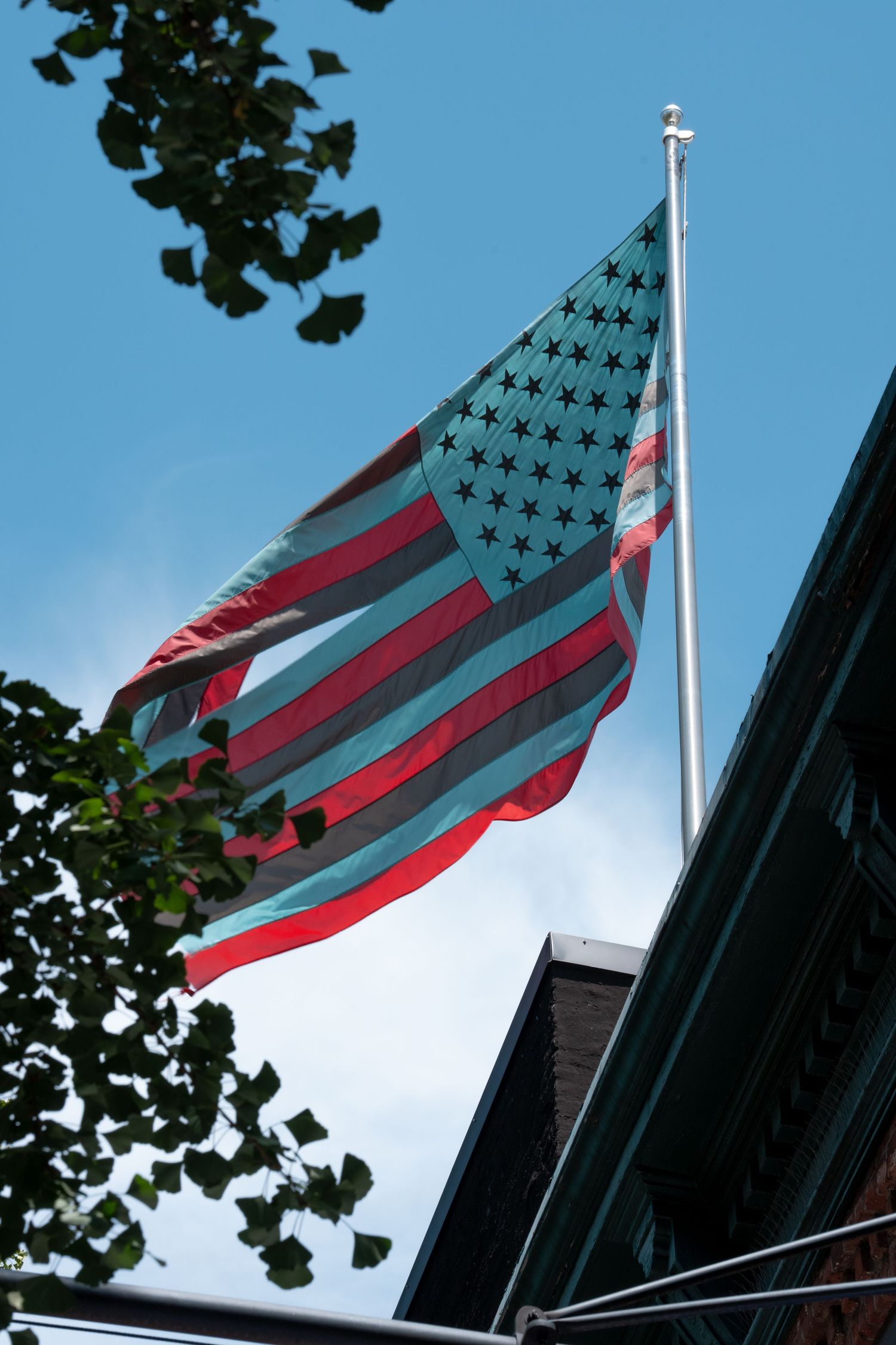 American flag waving against blue sky with tree leaves in foreground.