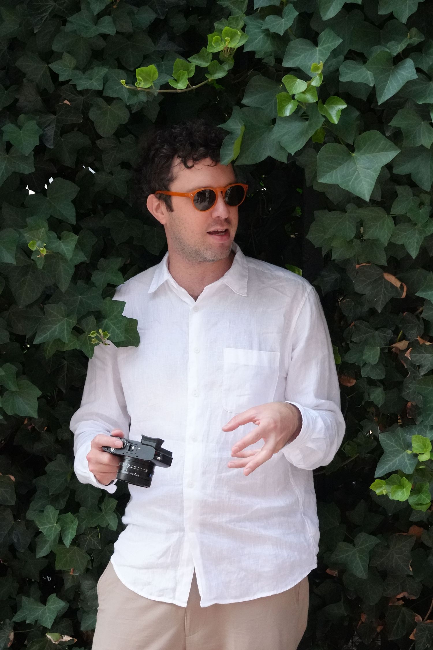 Person in white shirt and sunglasses posing against green ivy background.