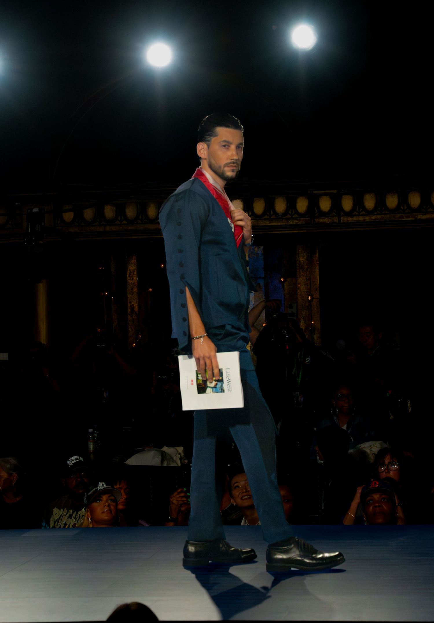 Model walks down a dark runway wearing navy blue outfit during fashion show.